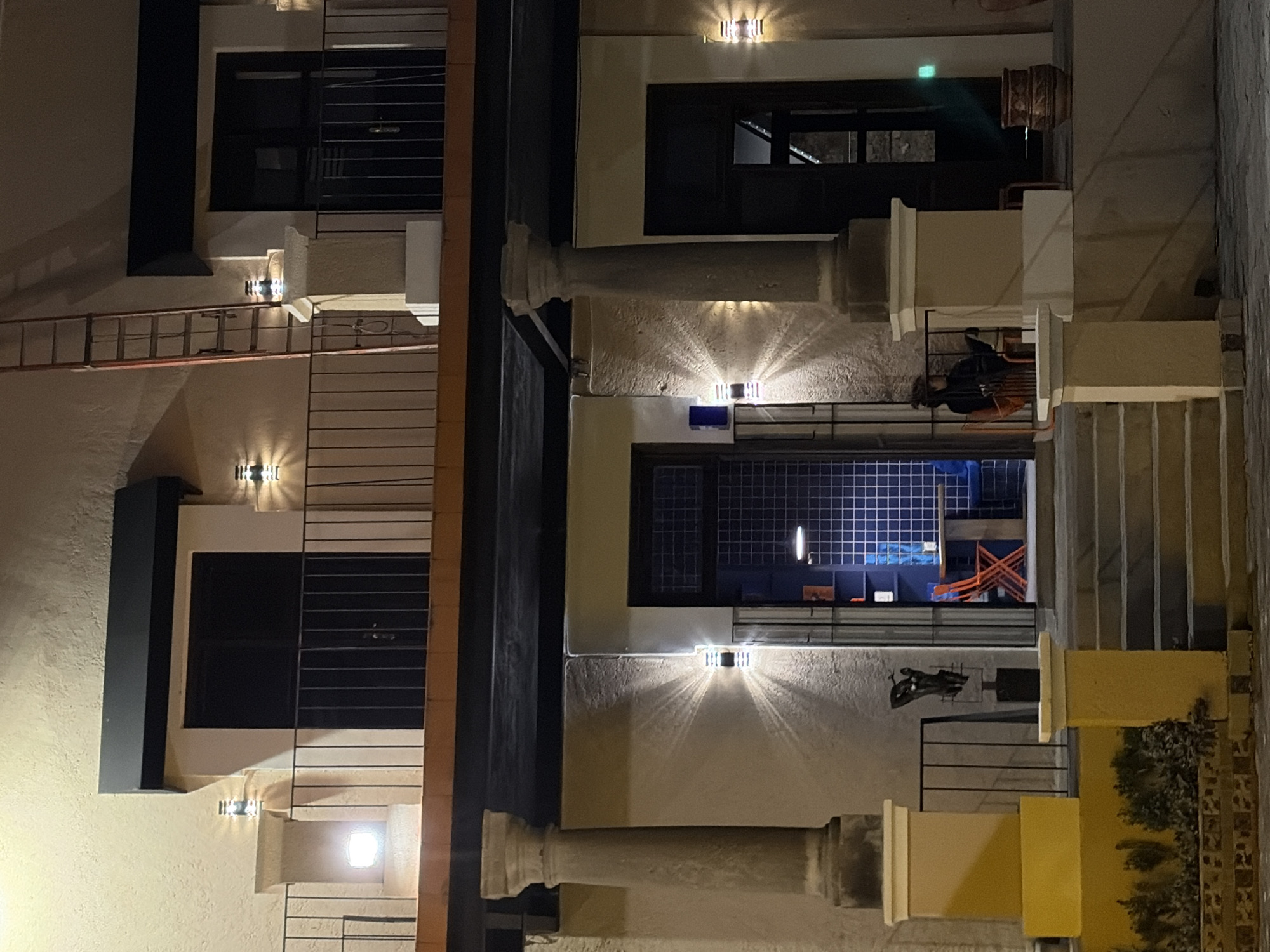 The image depicts a nighttime urban scene, likely in Puebla, Mexico, given the architectural style, showcasing a multistory building facade. The building features a mix of materials and textures, including light-colored stucco walls, dark window frames, and decorative lighting fixtures that cast geometric patterns on the walls. A variety of building elements are visible, such as ladders, stairs, balconies with metal railings, and cylindrical architectural features.
The presence of a dark figure on one of the balconies indicates human activity. The open window displays what appears to be an interior space illuminated by a blue light. The exterior of the building is illuminated by strategically placed light fixtures. The scene is captured from a perspective that emphasizes the verticality and intricate details of the building's construction.