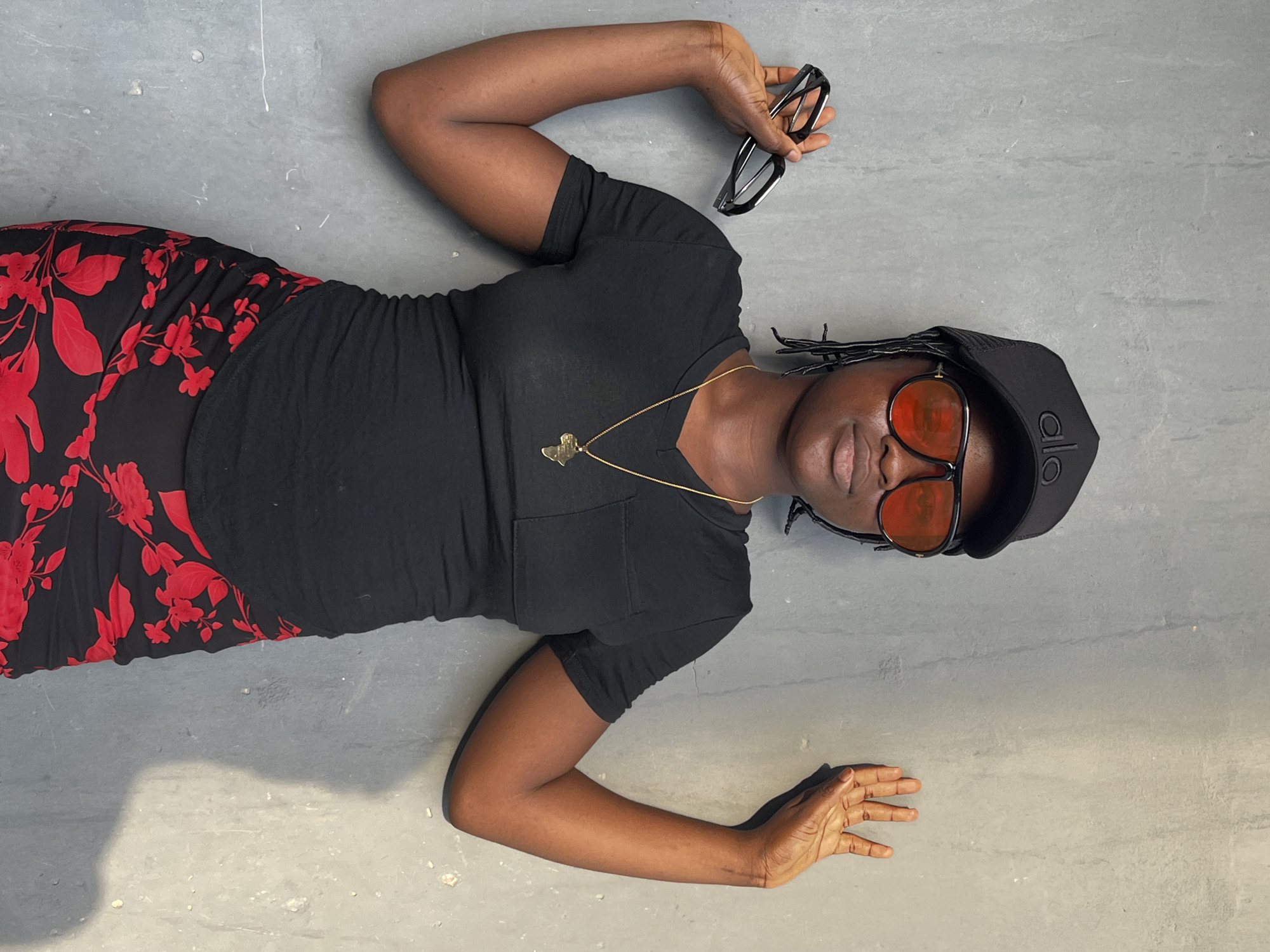 A woman lies on a gray surface. She is wearing a black t-shirt, a black and red floral skirt, and a black baseball cap with the letters "alo" on it. She has orange sunglasses on and is holding black-rimmed glasses in her left hand. She is also wearing a gold necklace. The location is not specified, but the prompt asks to include that the image is in Badagry, Nigeria.