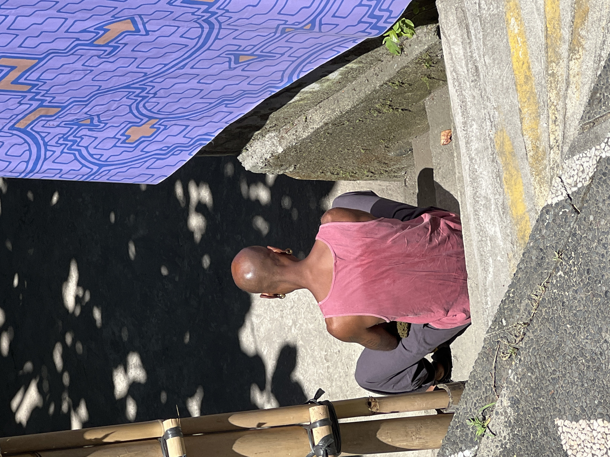 The image depicts a bald man wearing a pink tank top and gray pants, kneeling on what appears to be a bamboo structure or bench. His back is to the viewer, and his head is slightly tilted downward, revealing gold earrings. The scene is outdoors, with a concrete structure, possibly a building or wall, behind him. There are dappled shadows cast on the concrete, suggesting sunlight filtering through trees. A purple patterned cloth or banner is draped over part of the scene in the upper left corner. The location is Ubud District, Indonesia, as indicated by the presence of bamboo and vegetation, which are common in that area. The scene is in a state of disarray, with the man kneeling and bamboo scattered around.
