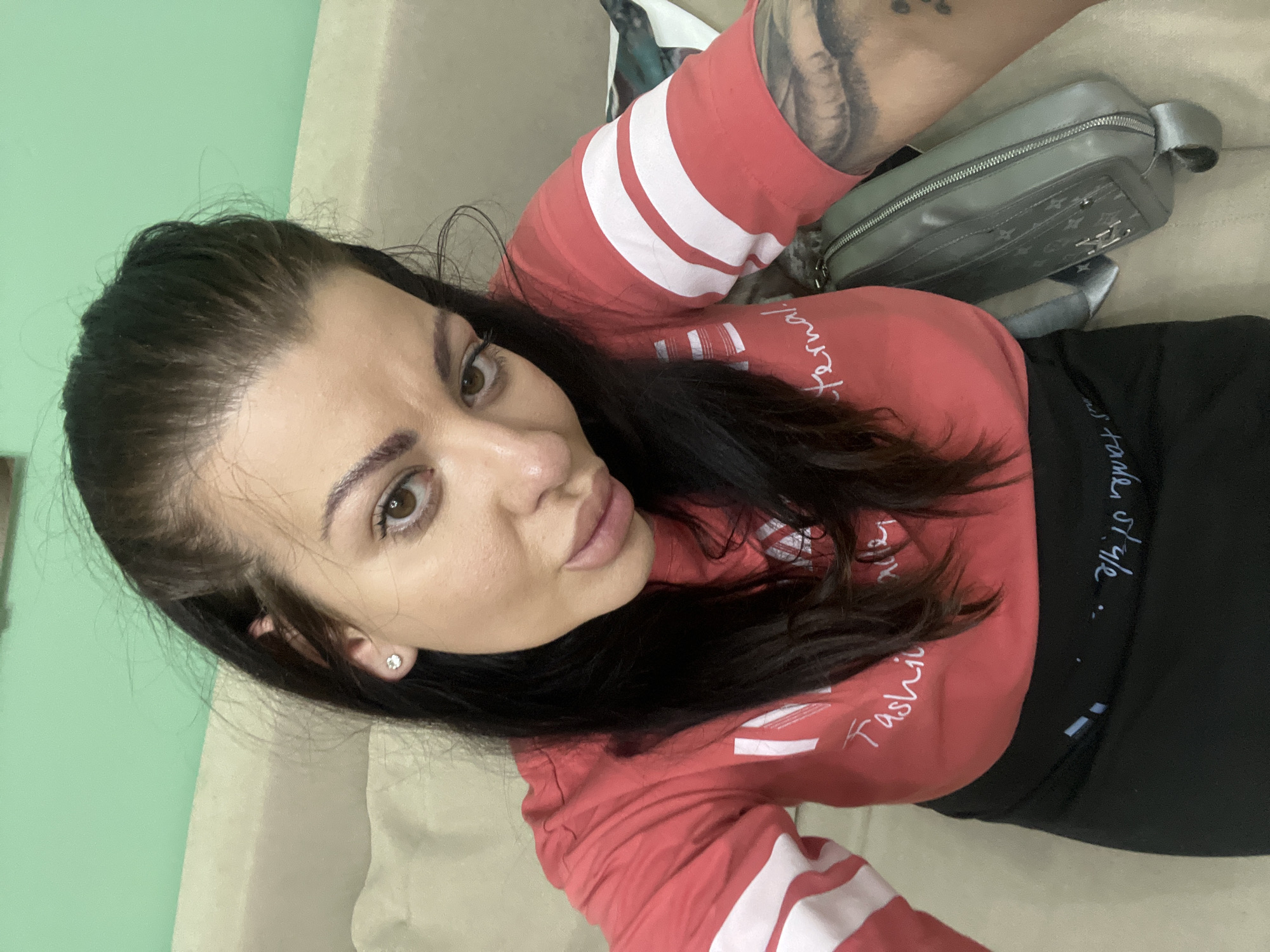 A young adult female with dark hair and brown eyes is taking a selfie from a seated position. She is wearing a red shirt with white stripes on the sleeves and a black skirt or pants. A grey purse with a patterned design is visible beside her. The background appears to be a couch and a mint green wall. The image was taken indoors, likely in a residential setting, and there is no indication of the specific location in Plovdiv, Bulgaria.