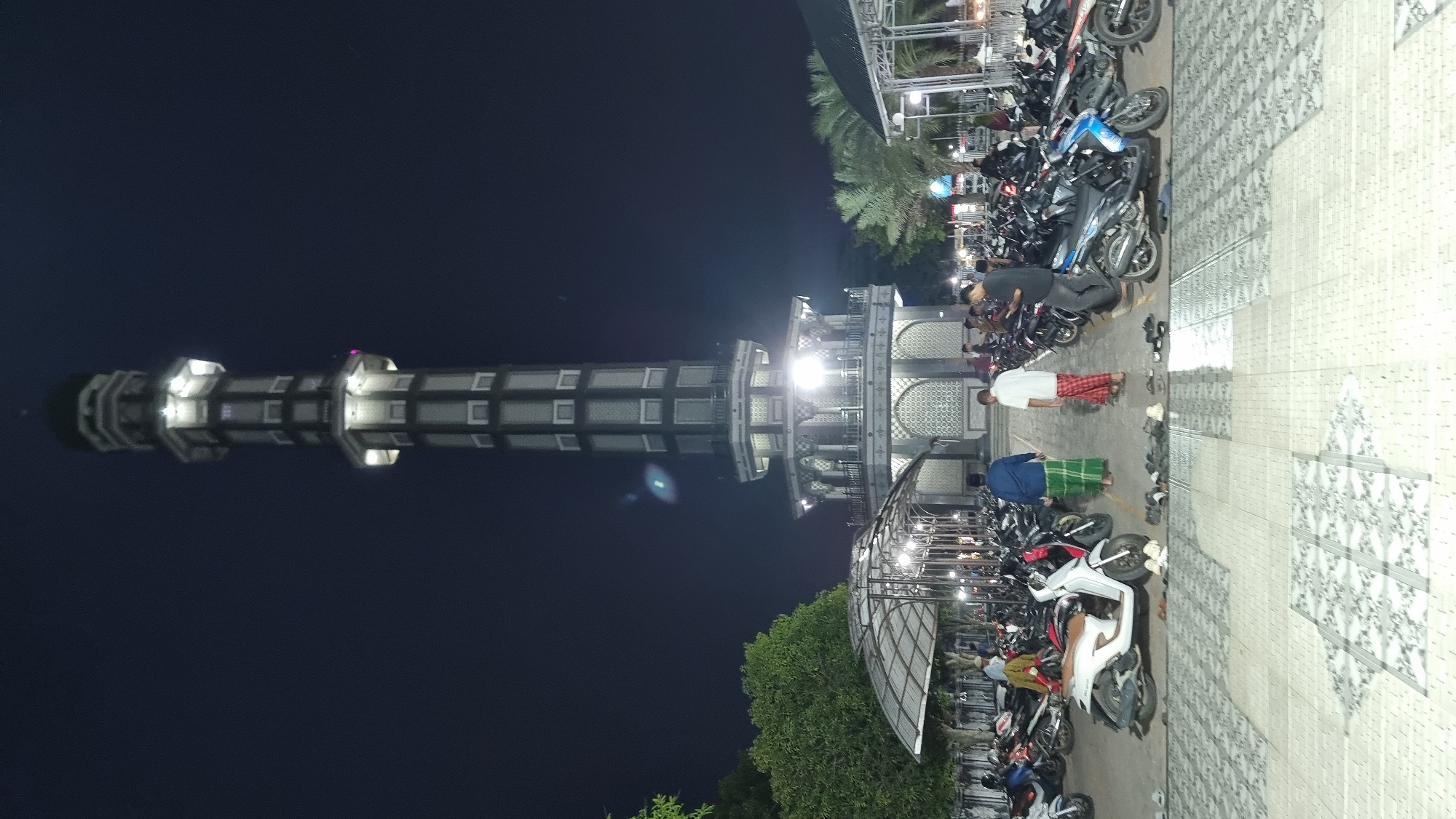 A vertical, outdoor, nighttime photograph shows a minaret of a mosque with a dark blue sky in the background. The minaret is tall and slender, with a dark patterned exterior and illuminated windows. It has a rounded top with a dark dome. Below the minaret is a wide, open area with a patterned floor. Numerous motorcycles are parked in rows across this area. Several people are walking in the foreground and mid-ground, some wearing traditional Islamic attire, such as sarongs. A covered structure with a transparent roof, supported by metal poles, is visible to the left. Palm trees and other foliage are scattered throughout the scene. The overall impression is of a mosque courtyard at night, with a significant number of parked motorcycles, suggesting it is a place of worship that attracts many visitors. The location is identified as Keudekruenggeukueh, Indonesia.