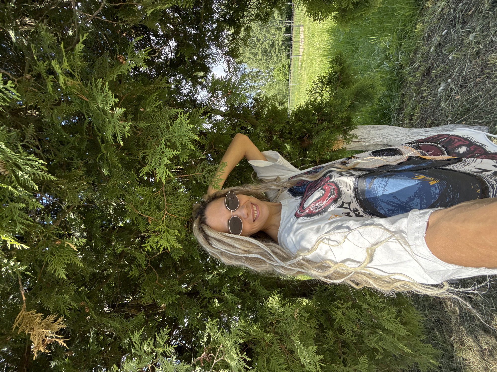 A woman with long, light blonde dreadlocks and round, dark-lensed sunglasses lies on her back, smiling brightly at the camera. Her head is positioned towards the upper part of the frame, surrounded by the dense, feathery green foliage of a coniferous tree or shrub, possibly a Thuja. She wears a white t-shirt featuring a large, colorful graphic design with abstract, swirling patterns in shades of blue, red, black, and white.
Bright sunlight filters through the tree's branches, indicating a clear, sunny day. Through a gap in the foreground foliage, a grassy area and a dark fence are visible in the background, suggesting an outdoor, possibly park-like setting. Her expression is joyful and relaxed. There is no visible text in the image. The scene is set in Rusanovo, Russia.