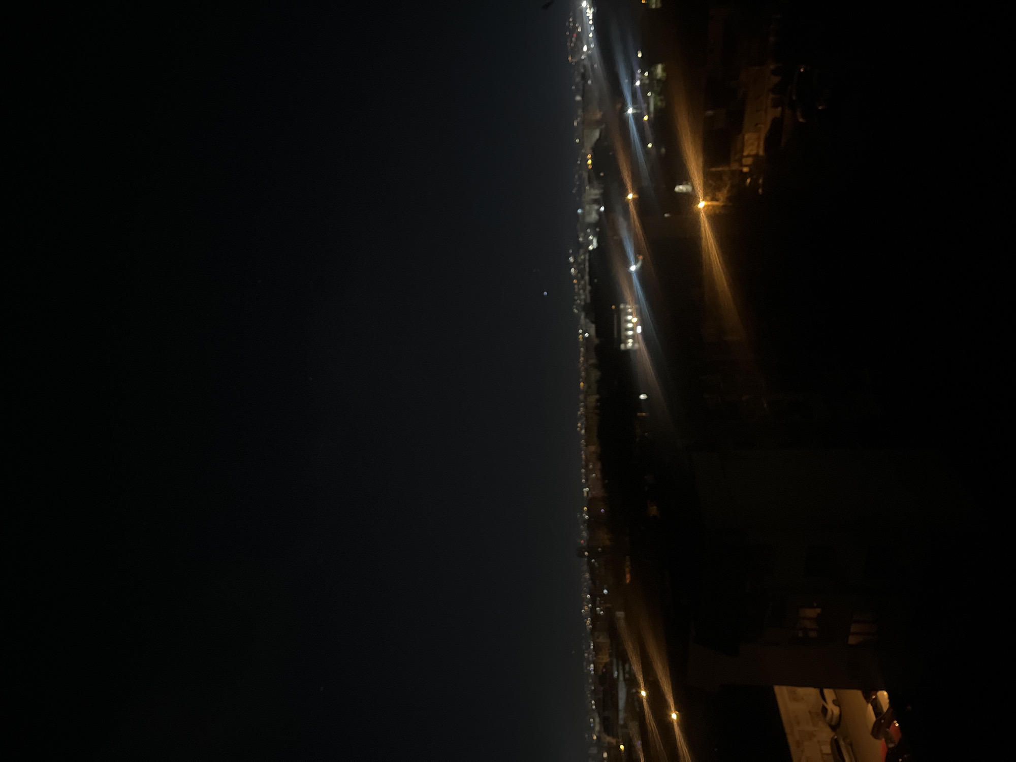 The image depicts a nighttime cityscape, likely taken from a high vantage point. The left side of the frame is dominated by a dark sky or body of water, with a few faint stars visible. The right side of the image shows a city illuminated by streetlights, creating streaks of yellow and white light. Some buildings and cars are also visible. Based on the presence of distinct orange street lighting and overall layout, the scene resembles a portion of the Sofia, Bulgaria urban environment, possibly the vicinity of the city.