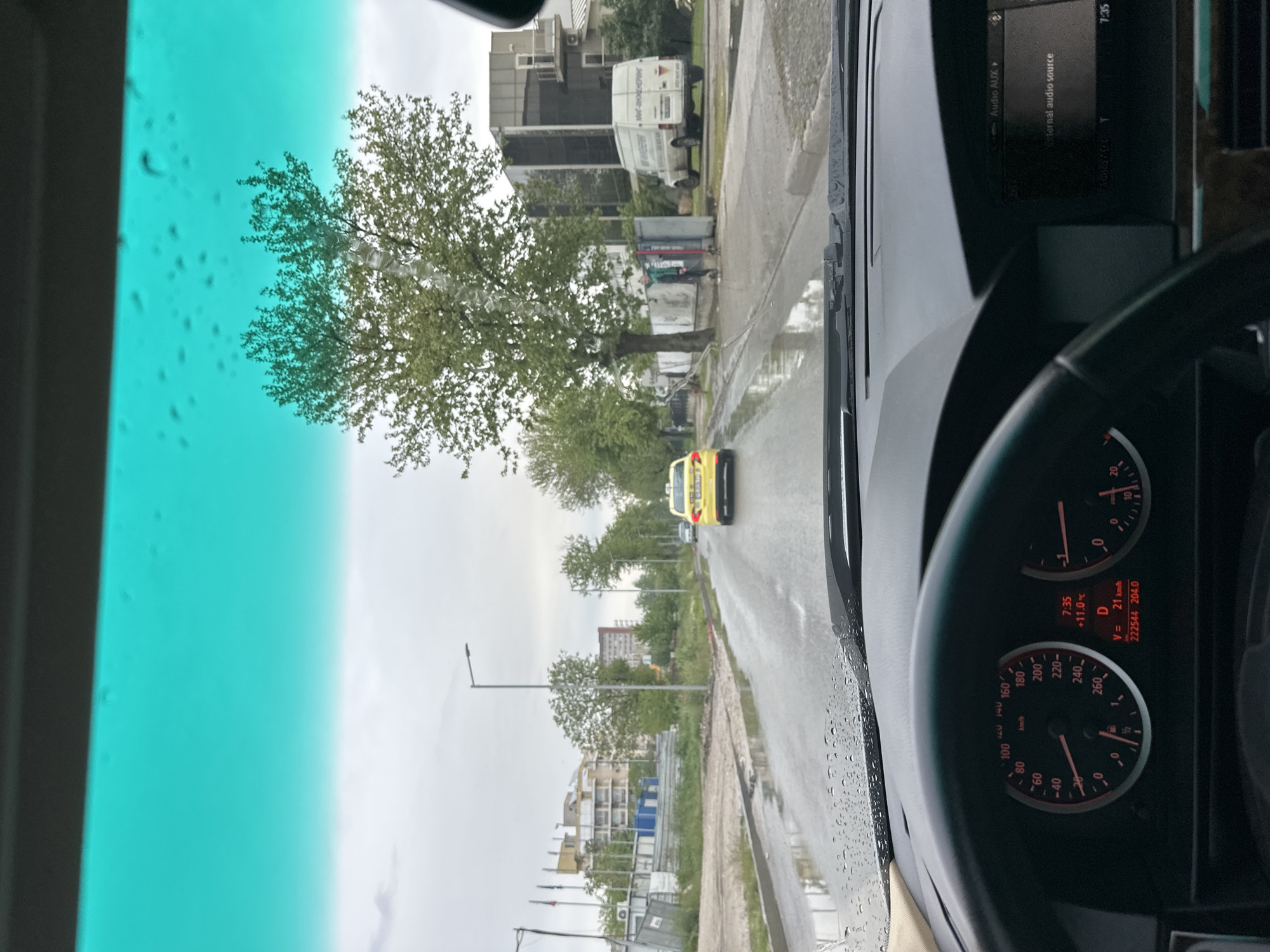 The image shows a view from inside a car on a wet road in what appears to be a city environment. The driver-side windshield is green-tinted and has raindrops on it. Through the windshield, there's a view of a tree-lined street with buildings in the distance. A yellow taxi is visible on the road ahead.
Inside the car, the dashboard, steering wheel, and speedometer are visible. The speedometer shows a speed of 21 km/h, and the dashboard displays a time of 7:35, with the temperature at +11.0°C. The odometer reading is 222544.
Based on the writing on the buildings, the location is Plovdiv, Bulgaria. One of the signs says "МИГ-ИНЖЕНЕРИНГ," which indicates the use of the Cyrillic alphabet, common in Bulgaria.