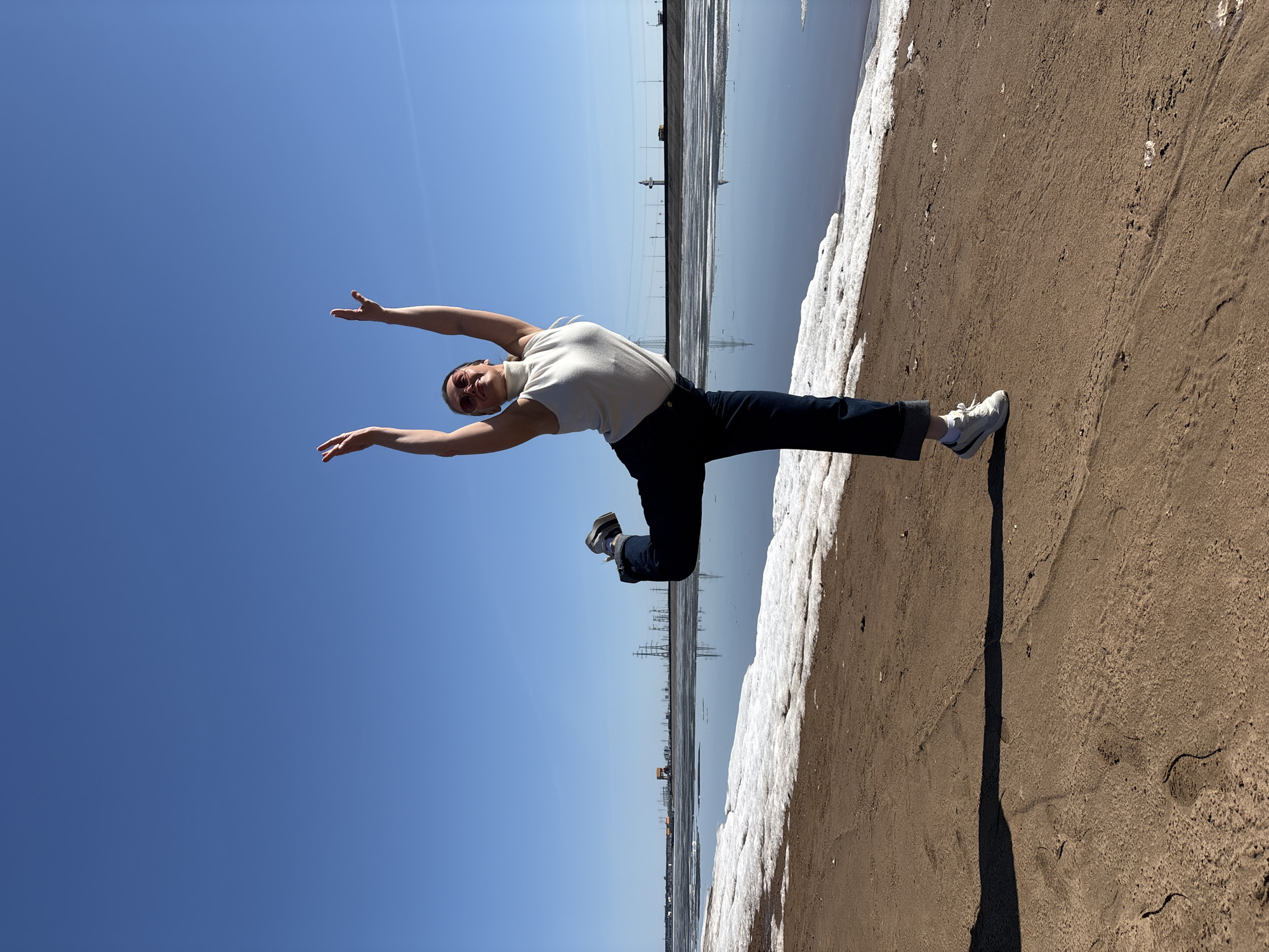 The image displays a dynamic outdoor scene with a person captured in a horizontal, mid-air pose against a bright blue sky. The subject, wearing a light-colored sleeveless top, dark pants rolled at the ankles, and white sneakers, has their arms outstretched and legs spread in a graceful, energetic leap or dance move. They are wearing sunglasses, and their expression is one of focus or joy.
The landscape is divided into distinct sections. The top portion of the frame is dominated by a vast, clear blue sky. Below this, a calm body of water stretches across the mid-section, reflecting the blue sky and distant industrial structures, including power lines and towers, which create vertical lines and shapes in the reflection. The lower part of the frame reveals a brown, sandy or muddy shoreline. A strip of melting white snow or ice runs along the edge where the ground meets the water.
The strong sunlight creates clear shadows on the sand, indicating a bright, sunny day, likely midday. The combination of sand and melting snow suggests a transitional season, such as early spring. The person's active pose and the clear, expansive environment evoke feelings of freedom and exhilaration. No text is visible within the image. The scene is set in Chaikovsky, Russia.