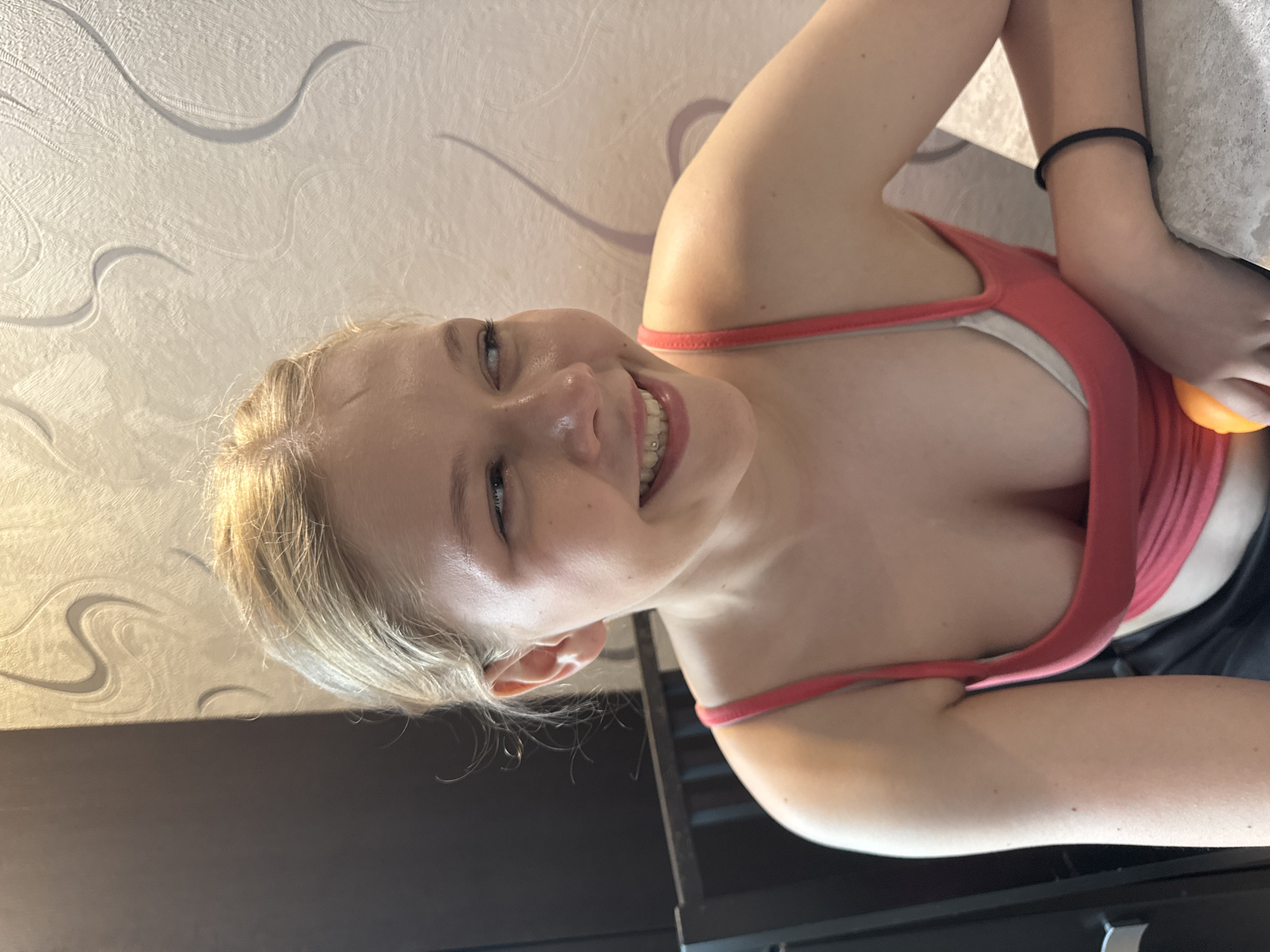 The image depicts a young Caucasian female, approximately from the chest up, in an indoor setting. She has blonde hair, fair skin, and is smiling, looking towards her right (viewer's left). Her mouth is open slightly, revealing teeth with a small, circular dental gem affixed to her upper left incisor. She wears a coral-red or orange-red thin-strapped tank top, beneath which a white undergarment or bra is visible. The low neckline of the tank top reveals cleavage. A plain black elastic hair tie is worn on her left wrist. Her left hand, partially obscured, holds a spherical orange fruit.
The background comprises two distinct vertical surfaces: a dark, matte, possibly wooden or laminate panel to the viewer's left, and a textured, light-colored wallpaper to the viewer's right. The wallpaper features an embossed or raised pattern of stylized, curvilinear swirls. Sunlight or bright artificial light illuminates the scene from the left, casting warm golden tones on parts of the wallpaper and creating subtle shadows within its texture. A dark, slatted back of a chair is partially visible behind her left shoulder. She rests her right arm on a light-colored, possibly concrete or stone, tabletop surface.
The specific location within the city of Perm, Russia, is not discernible from the visual information present in the image.