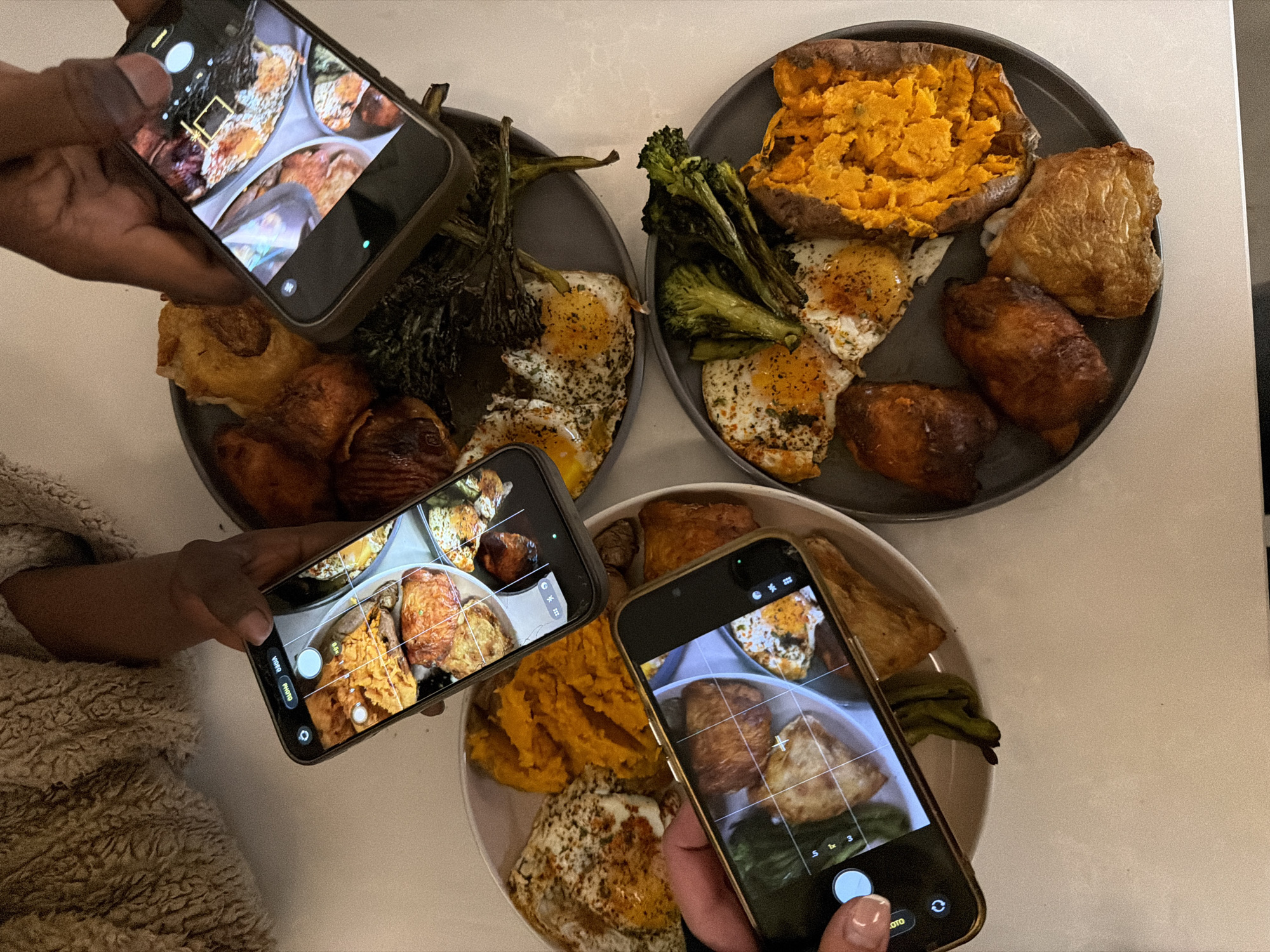 The image shows three plates of food being photographed by three different people using smartphones. The plates are placed on a light-colored surface, possibly a countertop or table. Each plate contains a variety of foods, including fried eggs, baked sweet potato, roasted vegetables (broccoli, other leafy greens), and some kind of meat (chicken). The people are positioned around the plates, holding their phones at different angles to capture images of the food. The phones have camera interfaces visible, suggesting they are actively taking photos.