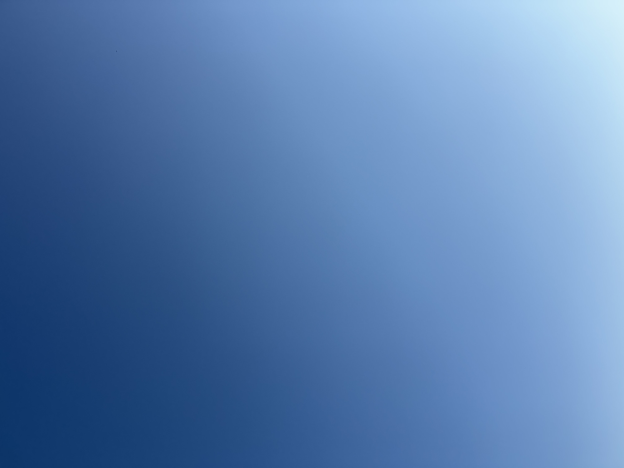 A clear, gradient blue sky with no discernible clouds. The sky transitions from a deep blue at the top of the image to a lighter, almost white hue at the bottom. A single, small black speck, possibly a bird or insect, is visible in the upper right quadrant of the image. The location is Atameken, Kazakhstan.