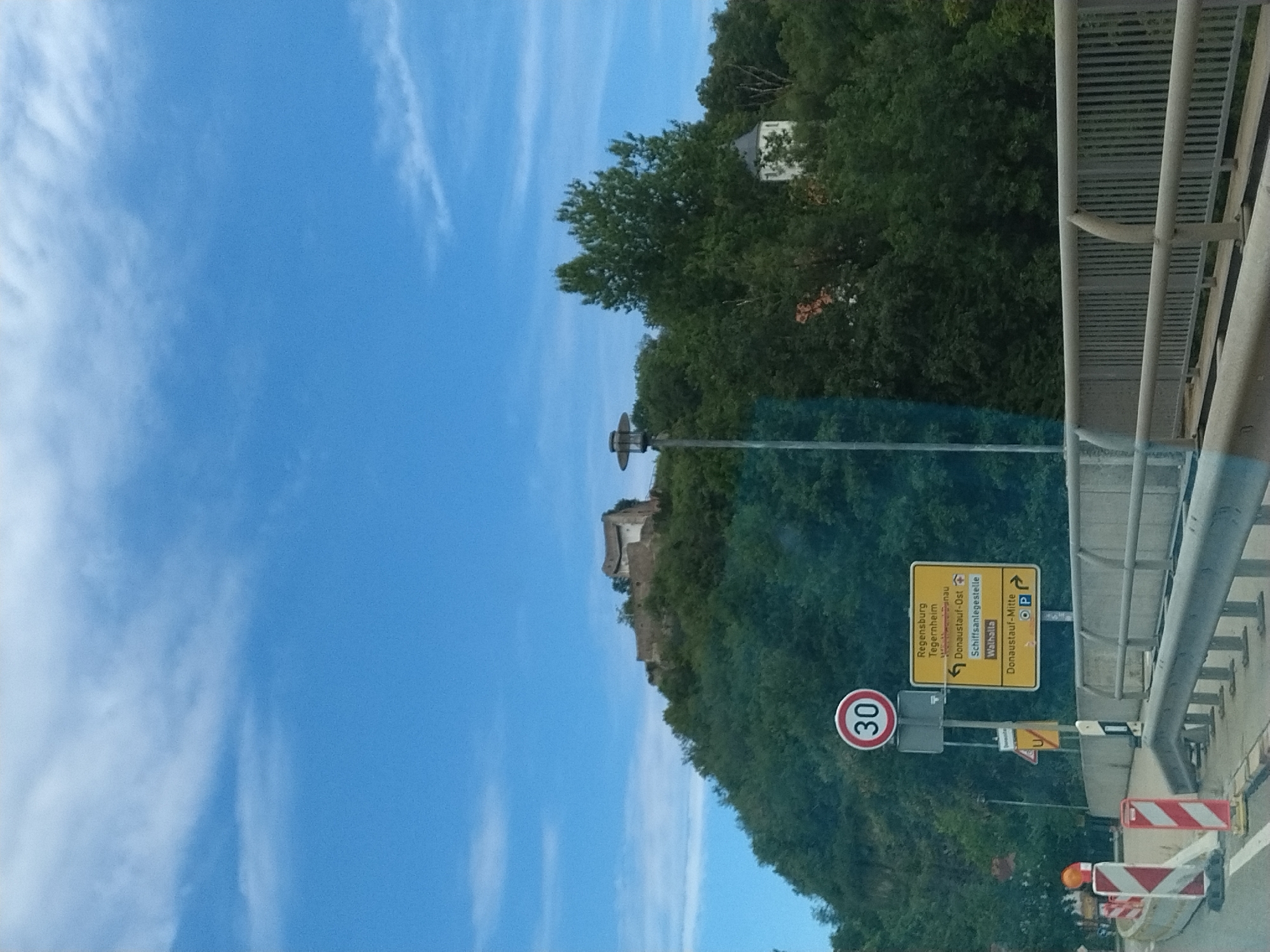 The image depicts a scenic view from a road in Donaustauf, Germany. A bridge with a metal railing runs across the foreground, with traffic cones and barriers indicating a construction or lane closure. Beyond the bridge, a lush green hill rises, topped by the ruins of a castle. A speed limit sign displaying "30" is visible on the left, next to a yellow directional sign indicating directions to Regensburg, Tegernheim, Donaustauf-Ost, Schiffanslegestelle, Walhalla, and Donaustauf-Mitte. A tall lamppost stands on the left side of the hill. The sky above is a clear blue with scattered white clouds.