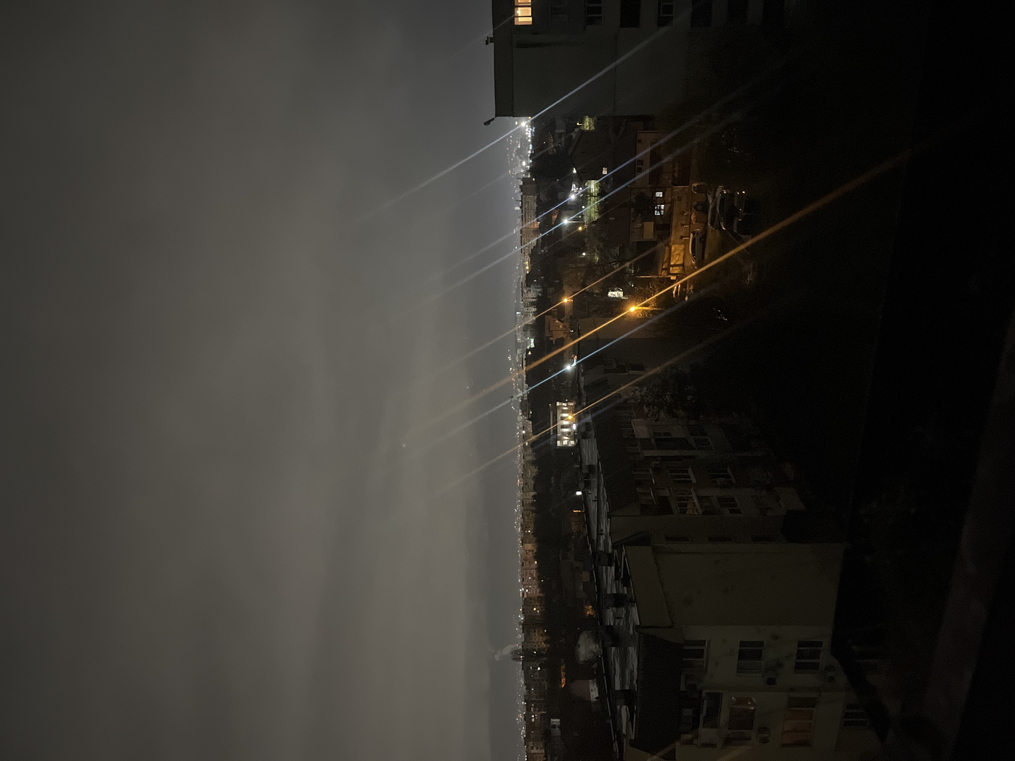 Here is a detailed description of the image:
The image shows a nighttime view of Sofia, Bulgaria. The scene is taken from a high vantage point, likely a balcony or rooftop, offering a panoramic view of the city stretching into the distance under a cloudy or foggy sky.
In the foreground, the corner of a building is visible on the right, its structure partially obscured by darkness. Moving towards the left, the city comes into view, illuminated by scattered lights from buildings and street lamps. The lighting creates streaks that cut through the sky.
The city appears densely built, with buildings of varying heights clustered together. The lights suggest active streets and inhabited residences. In the distance, the lights fade as the city stretches towards the horizon, blending into the darkness and overcast sky. The sky itself is a uniform dark gray, suggesting an evening or night with cloud cover.