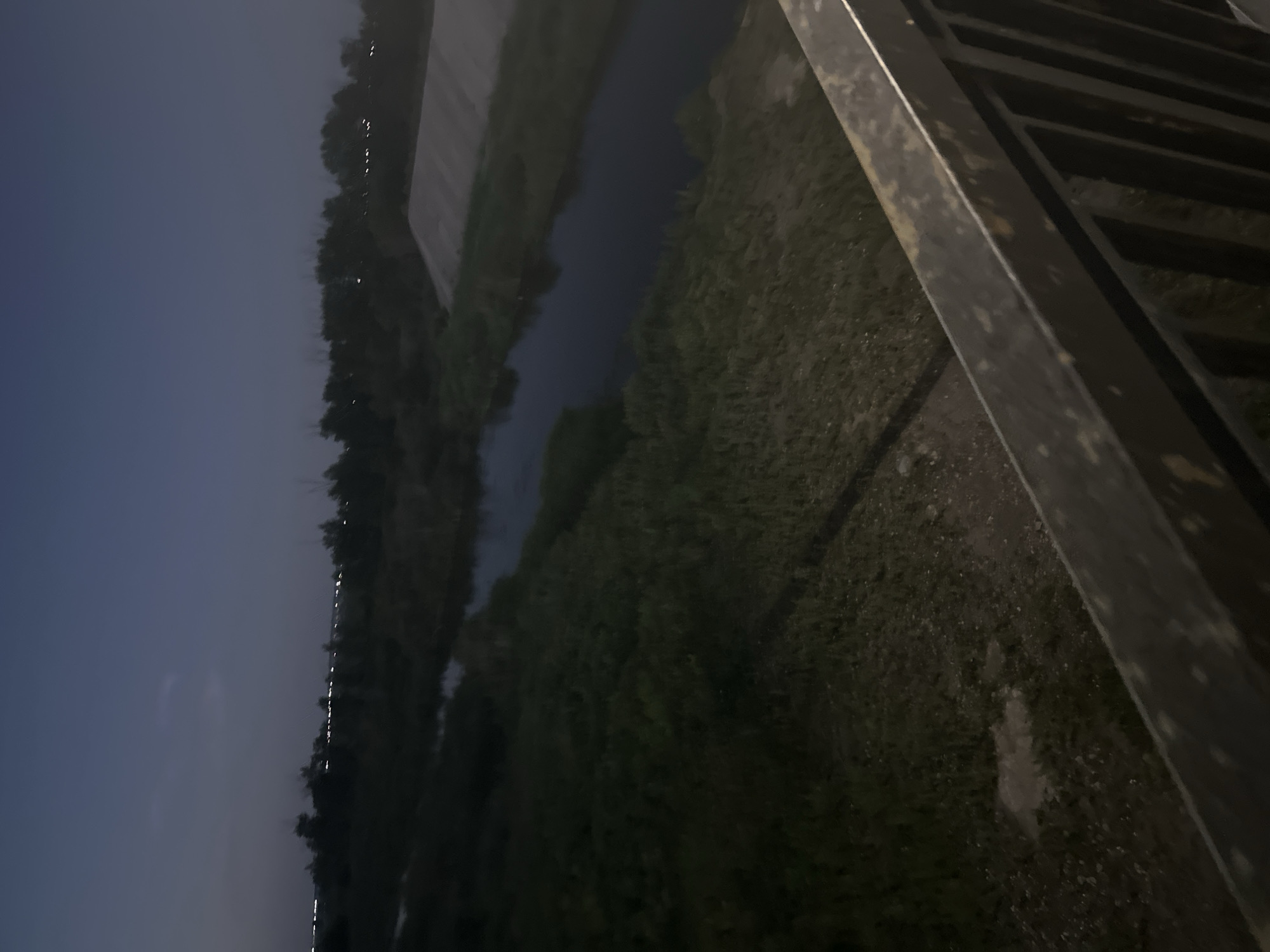 The image depicts a twilight view over a waterway in Kostanay, Kazakhstan. In the foreground, a dark metal railing with specks of rust is visible, suggesting a bridge or elevated walkway. Beyond the railing, a body of water, likely a river or canal, flows through a grassy embankment. The water reflects the dim sky, appearing dark blue. Dense vegetation lines the banks of the waterway, with trees silhouetted against the horizon. In the distance, faint lights indicate a developed area, possibly the outskirts of the city. The overall atmosphere is serene and quiet, with the fading light creating a dusky ambiance. There are no people or animals visible in the scene.