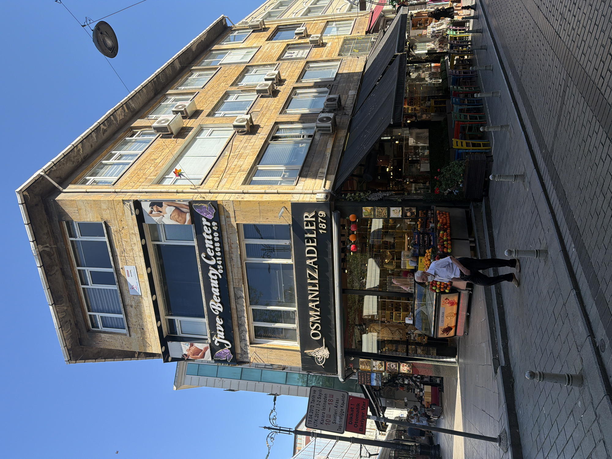 This media file depicts a street scene in Istanbul, Turkey, likely during the daytime given the clear blue sky and bright sunlight. The primary subject is a multi-story building with a facade of light-colored bricks. Several windows are visible, some with blinds, and numerous air conditioning units are mounted on the exterior.
On the ground floor of the building, there are storefronts with awnings. One prominent sign reads "Juve Beauty Center" with a phone number. Adjacent to it, another sign displays "OSMANLIZADELER 1879," suggesting a business or establishment with historical ties. The storefronts appear to house shops selling various goods, with displays of fruits and what looks like an ice cream freezer.
A person, an elderly man wearing a white shirt and dark pants, is walking on the tiled sidewalk. He is the only clearly visible person in the foreground. In the background, a few other figures are discernible, suggesting a moderately busy street.
A traffic sign is visible on the sidewalk, providing information in Turkish. It indicates a road closure for vehicular traffic between 10:00 and 18:00, referencing a decision dated 19.04.2012. It also warns of an automatic barrier. The street is paved with rectangular bricks, and decorative bollards line the sidewalk. The overall impression is of a typical urban commercial street in Istanbul on a sunny day.
