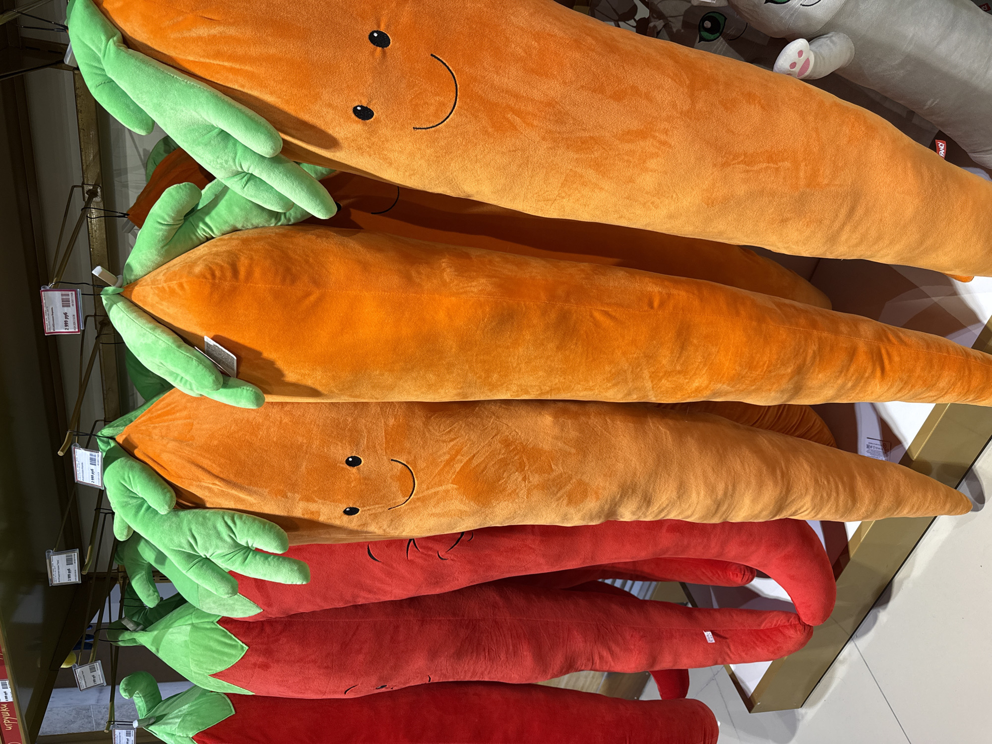 This image shows a stack of plush toys. The top three are orange carrot-shaped toys with green leafy tops and simple faces. Below them are two red chili pepper-shaped plush toys with green tops. The toys appear to be hanging on a display rack with price tags attached. The toys are presumably for sale. The photograph was likely taken in a shop in Moscow, Russia, based on a partially visible price tag.