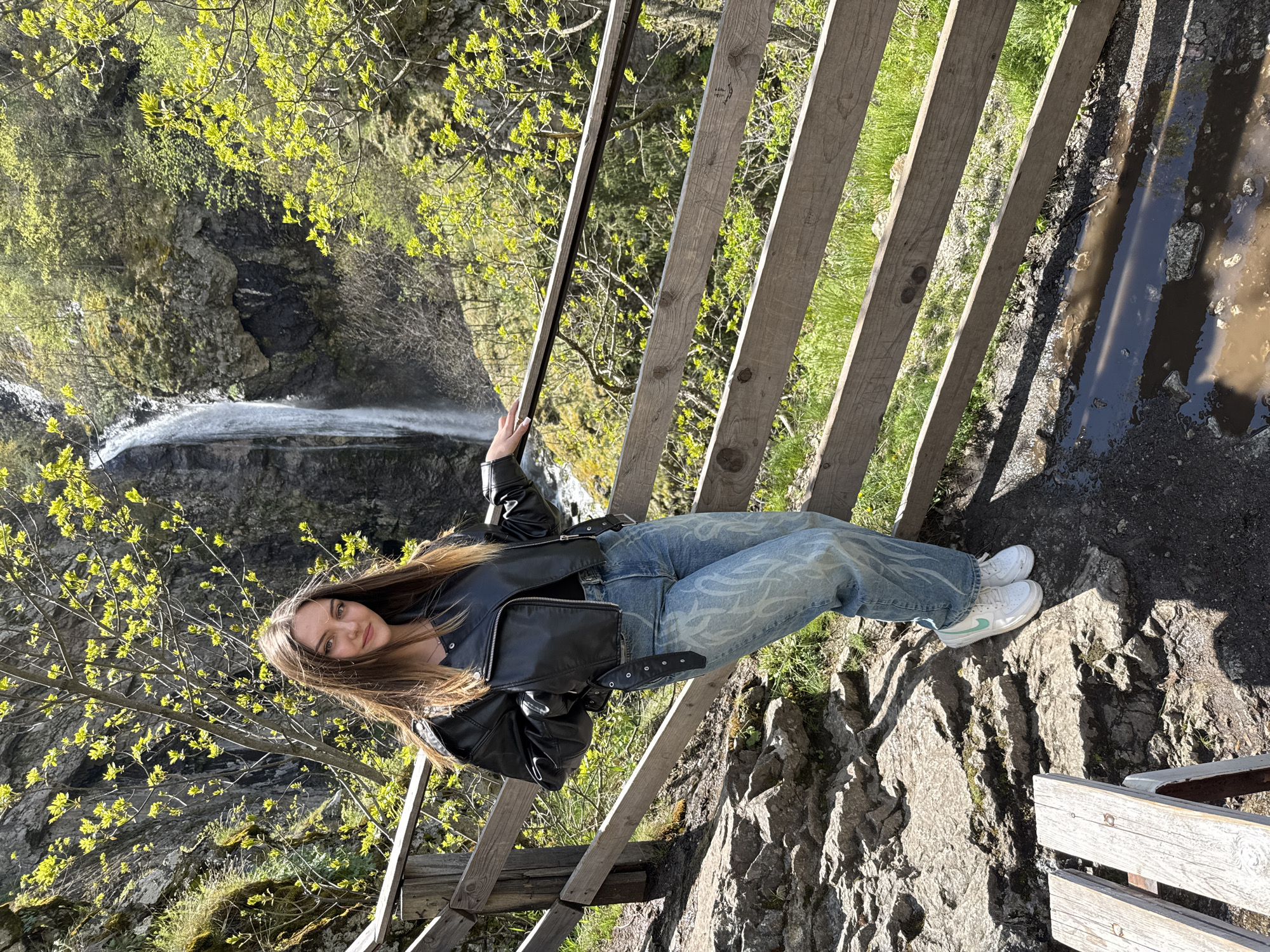A young woman poses on a wooden railing overlooking a waterfall in Ovchartsi, Bulgaria. She wears a black leather jacket, patterned jeans, and white sneakers. The waterfall is visible in the background, surrounded by trees and rocks. A puddle is to the right of the railing.