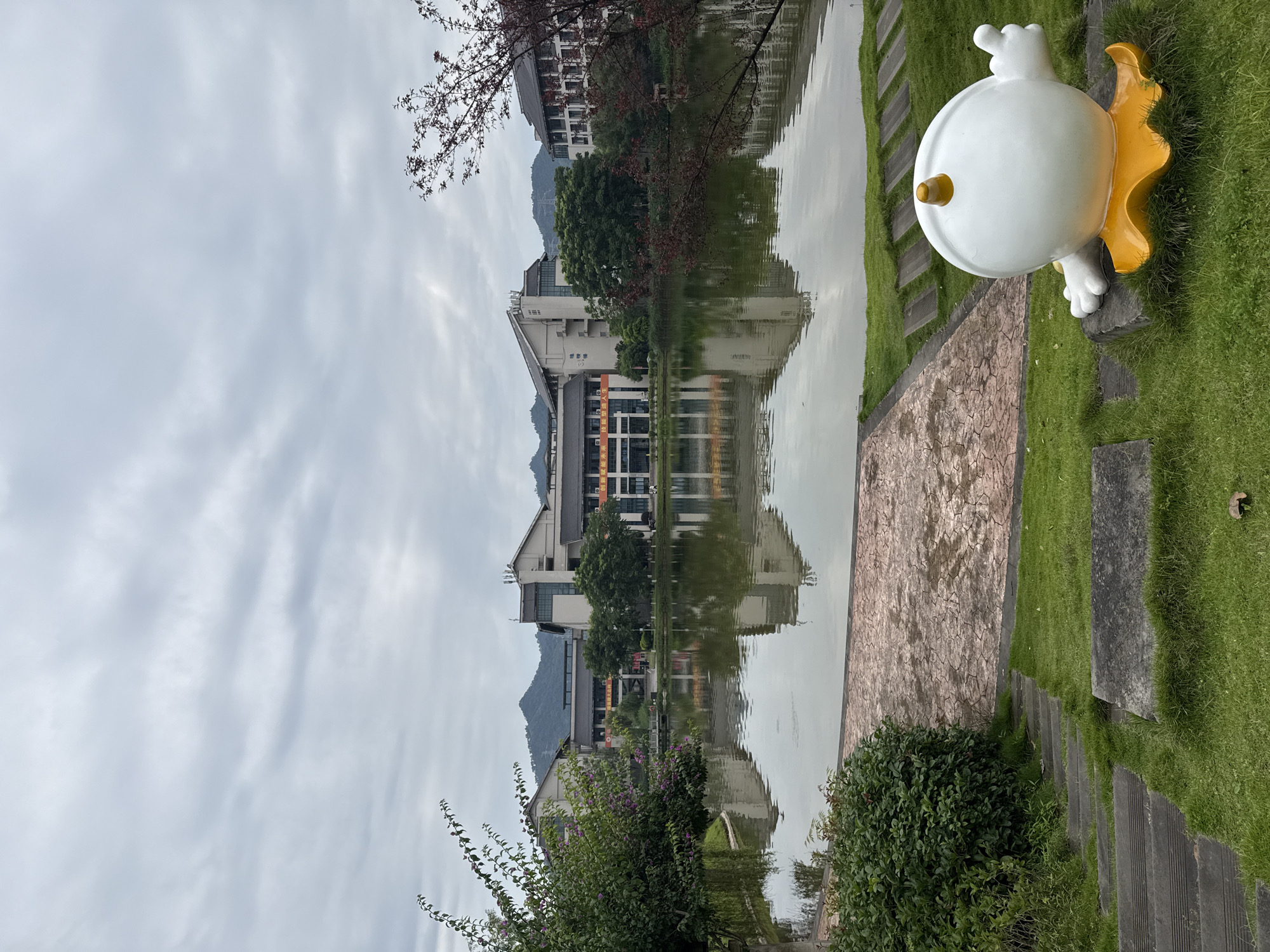 The image depicts a scenic view of a man-made lake with a multi-story building complex in the background. The building features a modern architectural style with a mix of white and gray facades, numerous windows, and some visible signage. Mountains are visible in the distance beyond the buildings. The lake's surface reflects the sky and the surrounding architecture. In the foreground, a white, cartoonish sculpture with a yellow base and horns sits on a stone base amidst green grass. A pathway of gray stones and an area of cracked-patterned stone pavement are also visible on the grass. Steps made of wood and stone lead down to the left of the frame. The sky is overcast with diffused light. The scene is likely located in the city of Jiangjin, China, given the context of the provided crops suggesting this location.