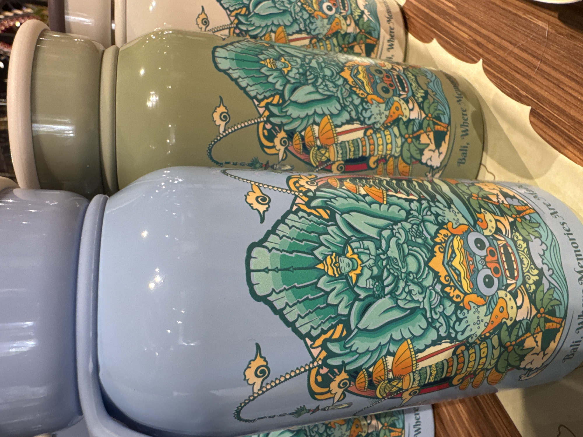 Three ceramic mugs are stacked horizontally, each bearing a similar design dominated by a stylized, green and orange figure. The text "Bali, Where Memories Are Made" is visible on the mugs. The mugs are light blue and green. The scene takes place in Kuta Selatan, Indonesia, suggested by the text on the mugs. The mugs are on a table made of wood and a white corrugated plastic placemat.