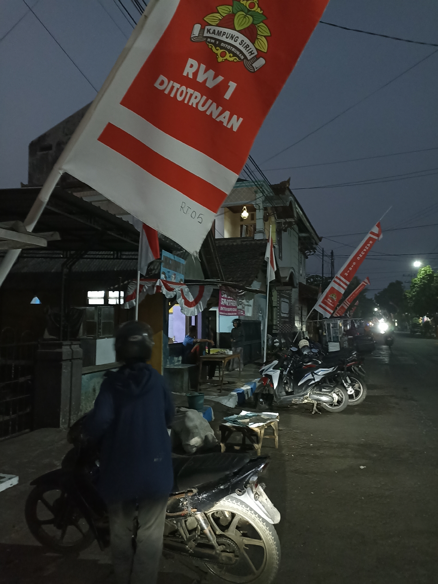 An outdoor, low-light scene depicts a street in Ditotrunan, Indonesia, during dusk or early evening. A prominent red and white flag with text in white is visible in the foreground, angled downwards, with "RW 1 DITOTRUNAN" and "KAMPUNG SIRIH" legible. To the left, a person wearing a dark hooded jacket and light-colored pants stands near a dark-colored motorcycle. Behind them, a covered structure with some seating is visible. Further down the street, several other motorcycles are parked. A building with a traditional Indonesian architectural style stands on the right side of the street. Trees line the street in the background, and streetlights are illuminated. The ground appears to be asphalt or dirt.