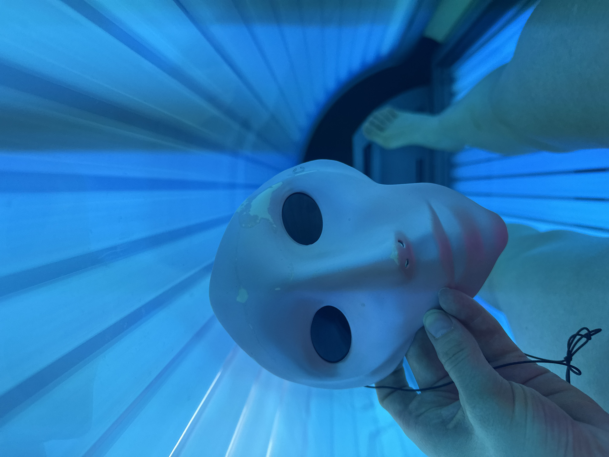A person is lying inside a tanning bed, holding a protective eye mask. The tanning bed emits a blue light. The scene is not identifiable as being in Saint Petersburg, Russia, and there are no objects present suggesting this location.