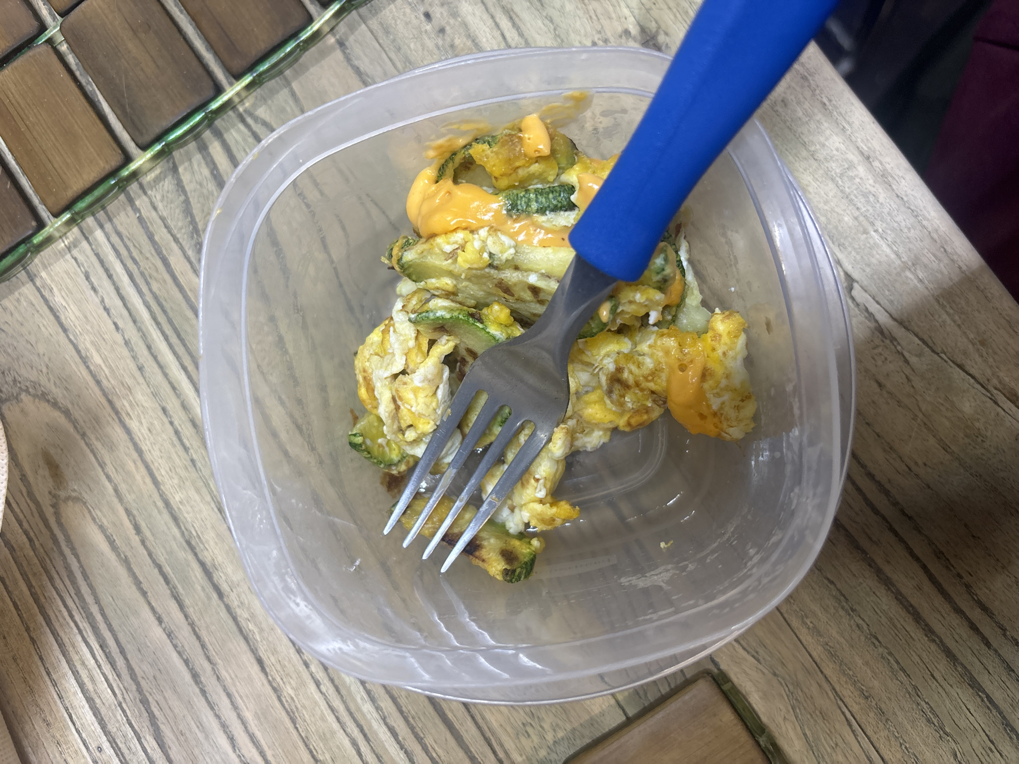 A close-up, high-angle shot shows a clear plastic food container filled with a mixture of scrambled eggs, sliced zucchini, and a creamy orange sauce, possibly cheese sauce. A blue-handled fork is inserted into the food. The container sits on a table or surface with a wood-grain pattern, alongside a section of a decorative table with a mosaic of small wooden tiles. There is no indication of the city of Buenavista, Mexico.