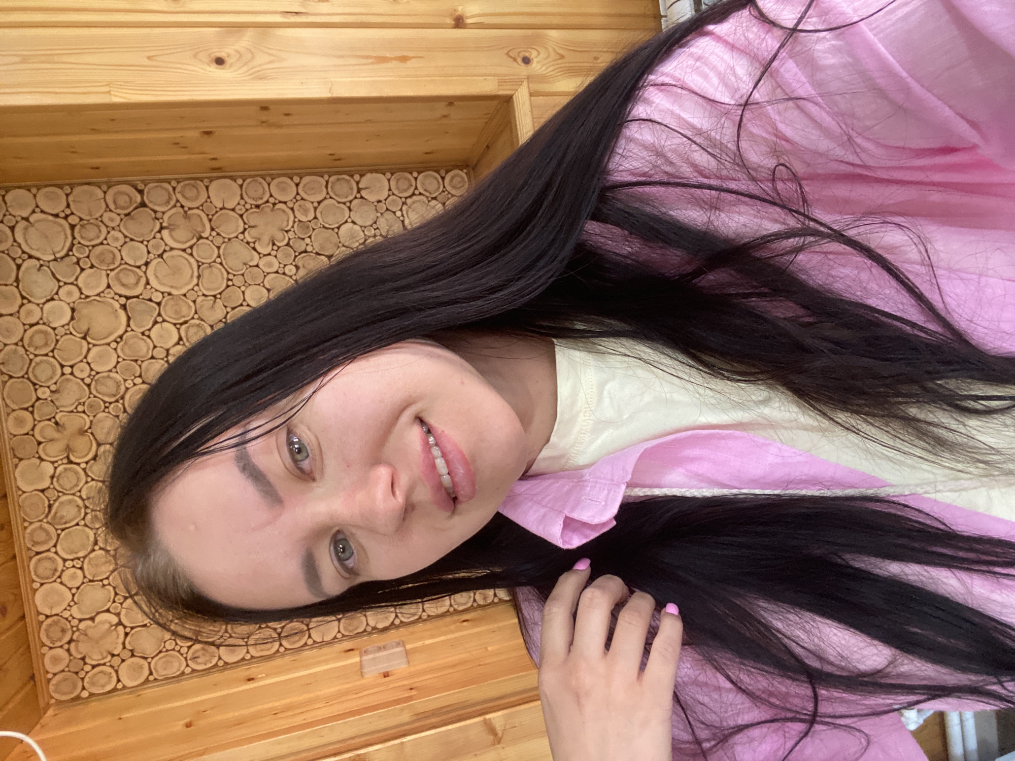 A young Caucasian woman with long, dark hair and light-colored eyes is looking at the camera and smiling. She is wearing a light yellow t-shirt and an open pink collared shirt. Her hand is raised to her hair, and her fingernails are painted pink. The background consists of light brown wooden walls with a decorative pattern of circular wood slices. The scene appears to be indoors, possibly in a room with a rustic aesthetic. There is no indication of the city of Anninsk, Russia within the image itself.