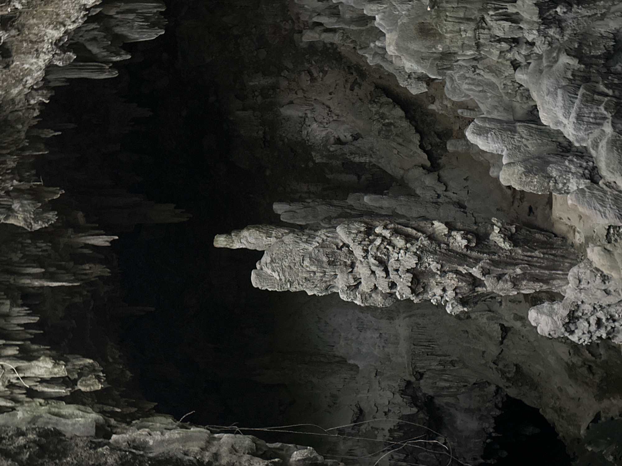 The image shows the interior of a cave, likely a tourist attraction, located in Xóm Khán, Vietnam. The cave features numerous stalactites hanging from the ceiling and stalagmites rising from the floor, creating a dramatic and natural landscape. A prominent, tall stalagmite stands in the center of the frame, surrounded by smaller formations. The lighting is dim, suggesting the natural ambiance of the cave, with shadows accentuating the textures and shapes of the rock formations. No people or animals are visible in the image. The overall environment is stony and cavernous.