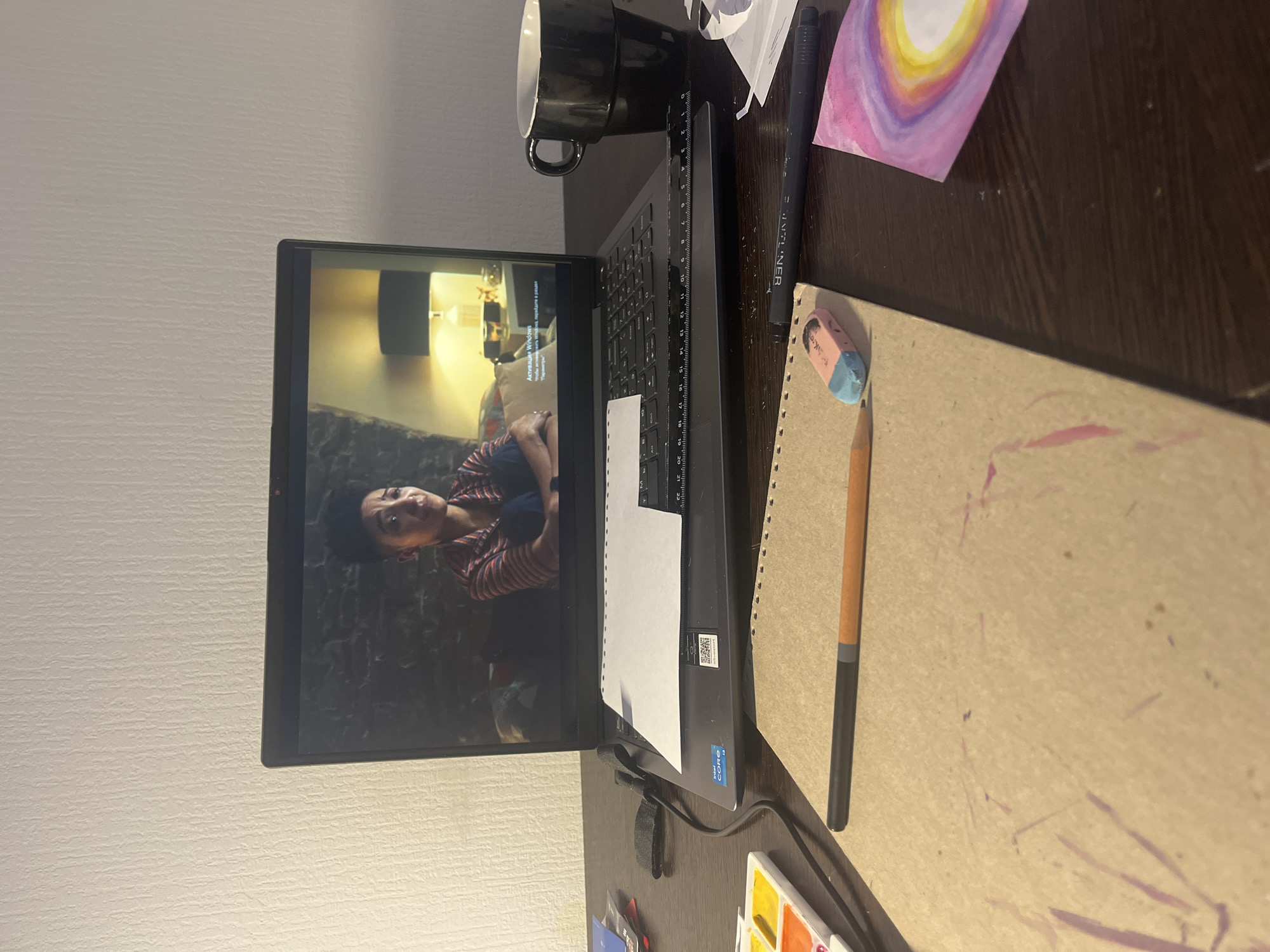 The image depicts a desk setting with a laptop as the central object. The screen displays a video featuring a person with short, dark hair, wearing a striped shirt and sitting in a room with a stone-like wall and warm lighting. A black mug rests to the left of the laptop, and to the right, there is a drawing pad with a pencil, eraser, and a black pen. A painting with colorful concentric circles is also present. A small watercolor palette can be seen at the bottom. The scene is indoors, but there's no specific indication in the image that it's in Saint Petersburg, Russia.