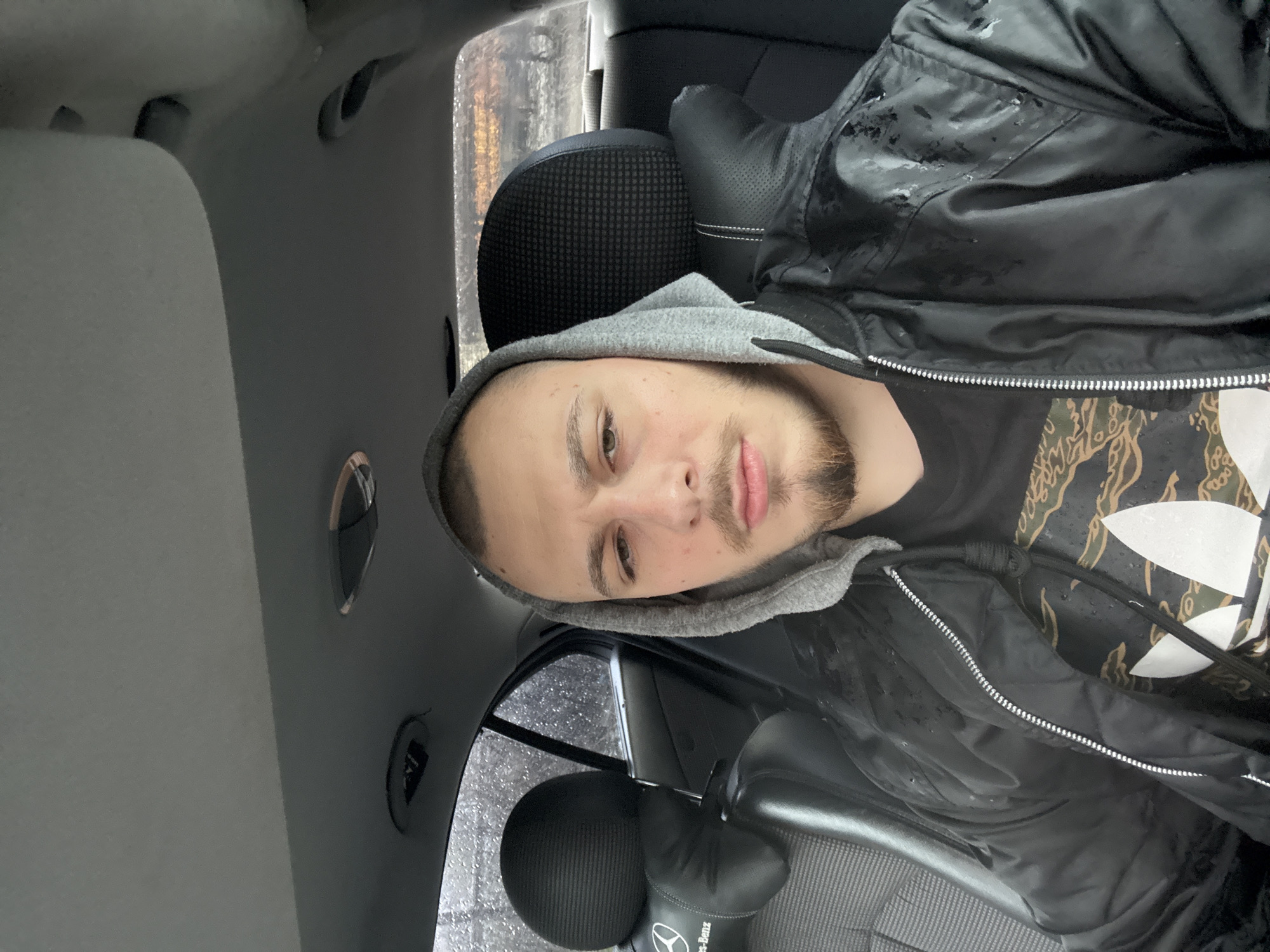 A man is seated inside a car, possibly in Stara Zagora, Bulgaria, given the query context. The car's interior is visible, including the ceiling, a headrest, a seat, and the side window. The man is wearing a grey hoodie under a black jacket over a dark patterned t-shirt. The exterior window shows a blurred street scene, possibly on a rainy day, with indistinct structures.