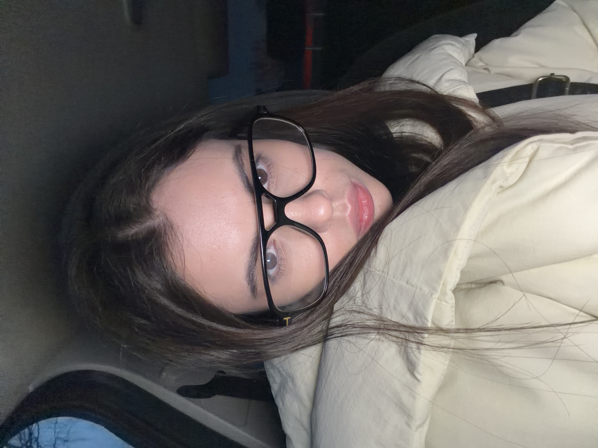 The image is an angled, close-up portrait of a young person, likely female, with long, dark brown hair. She is wearing black-framed glasses, and a small golden "T" logo is visible on the right temple piece of the frames. Her eyes, appearing light brown or hazel, are open and directed towards the camera. Her lips are slightly parted, displaying a natural, soft pink hue.
She is wearing a light-colored, possibly off-white or cream, padded jacket. A black belt with a gold buckle is visible resting on the jacket on her right side. The person appears to be reclining or lying down, given the perspective.
The background is largely dark and out of focus, suggesting a dimly lit indoor environment or the interior of a vehicle. In the lower left of the frame, there's a faint indication of a window or reflective surface revealing bare tree branches against a lighter, possibly overcast, sky, hinting at the time of day being late afternoon, evening, or early morning. The overall mood is calm and relaxed. Given the context of Chaikovsky, Russia, the padded jacket suggests cooler temperatures.