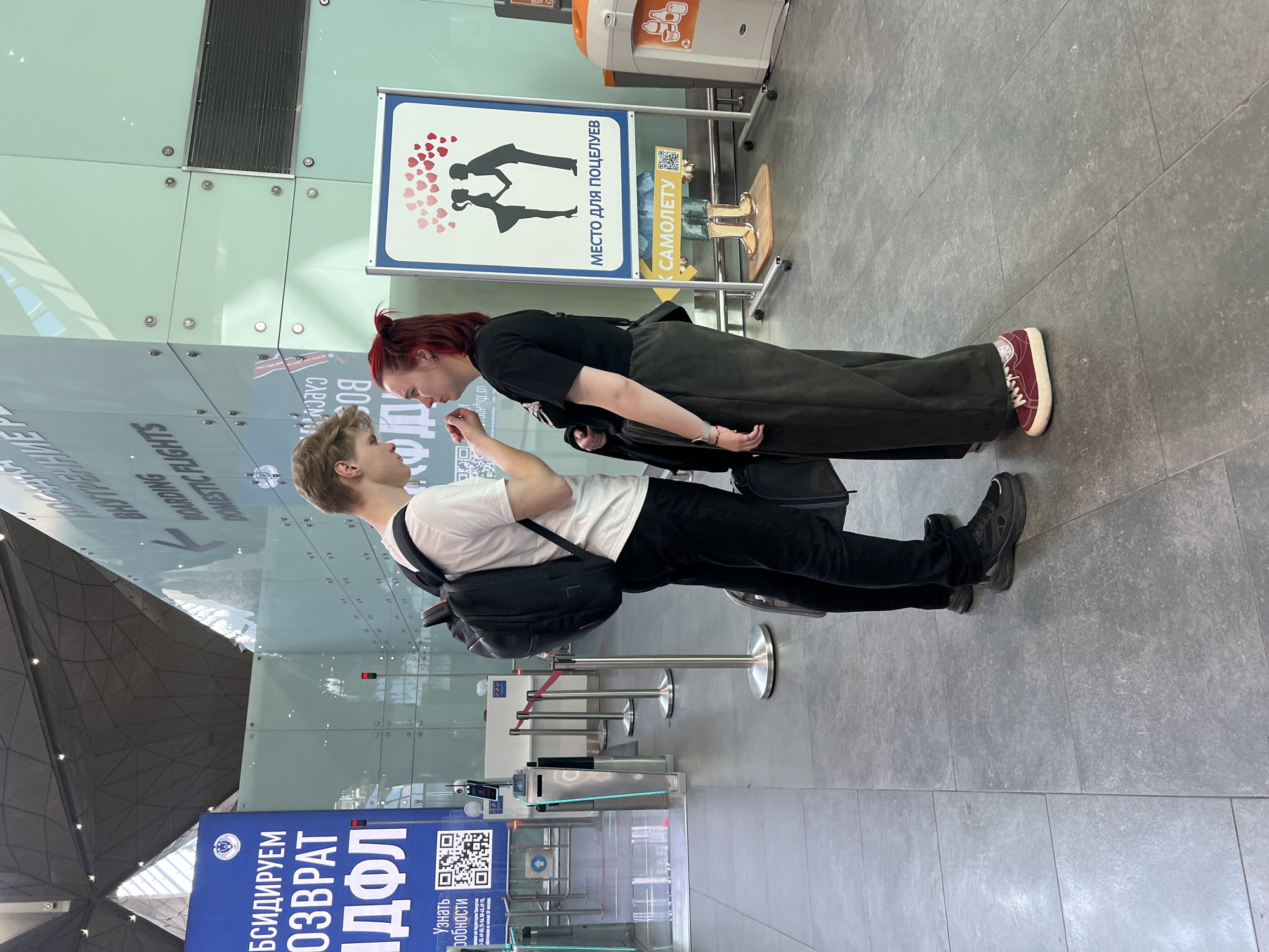 The image depicts a young couple interacting in what appears to be an airport terminal in Saint Petersburg, Russia. The couple is positioned face-to-face, with the woman leaning in towards the man, suggesting a moment of affection or connection. Both individuals are casually dressed; the woman has red hair tied up and is wearing a black top and dark, wide-leg trousers, complemented by maroon sneakers. The man has light-colored hair and is wearing a white t-shirt, black trousers, and black sneakers, with a backpack slung over his shoulder.
Behind them, a sign featuring silhouettes of a couple kissing surrounded by hearts is visible, with Russian text that translates to "Kissing spot." Another sign with yellow lettering that says "To the plane" is also present. The background shows the characteristic modern architecture of an airport, with glass walls, metallic structures, and signage indicating "DOMESTIC FLIGHTS." Several security gates and barriers are set up, typical of a departure area. A large blue banner with Cyrillic text and QR codes is also visible, possibly related to services or information. The lighting suggests it is daytime, and the overall atmosphere seems calm, with the focus on the intimate interaction between the couple amidst the functional environment of the airport.