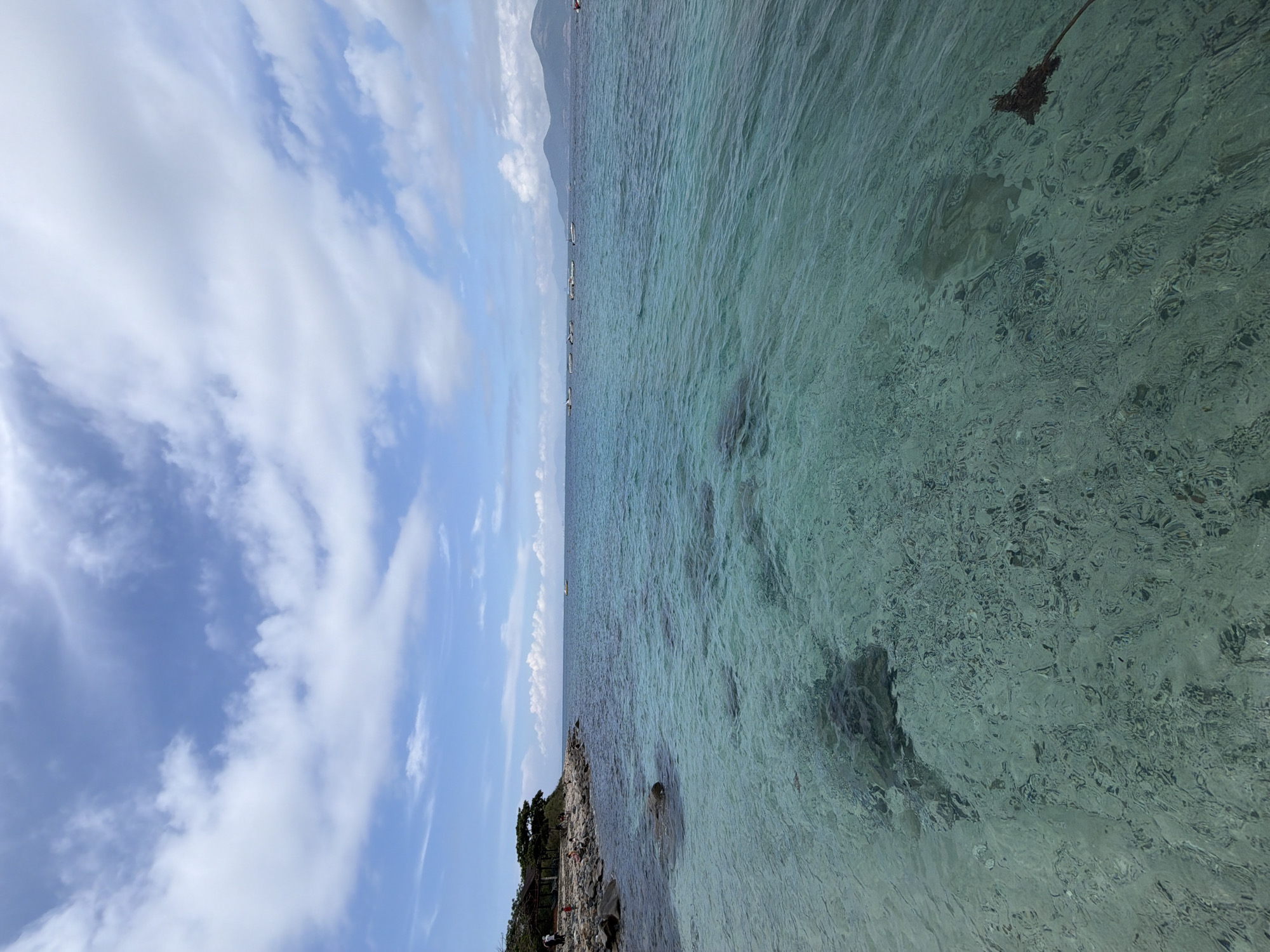 The image shows a coastal scene in Nha Trang Ward, Vietnam. The sky is blue with scattered white clouds. A body of clear, turquoise water stretches from the foreground to the horizon, where a mountain range is visible. Along the shoreline, there's a rocky area with sparse vegetation and a small structure, possibly a hut or gazebo. Several windsurfers/ sailboats are situated along the water near the mountain range. The image is taken from a slightly elevated perspective, capturing a wide expanse of the ocean and sky. The water is clear enough to see dark patches, which appear to be coral reefs or underwater vegetation.