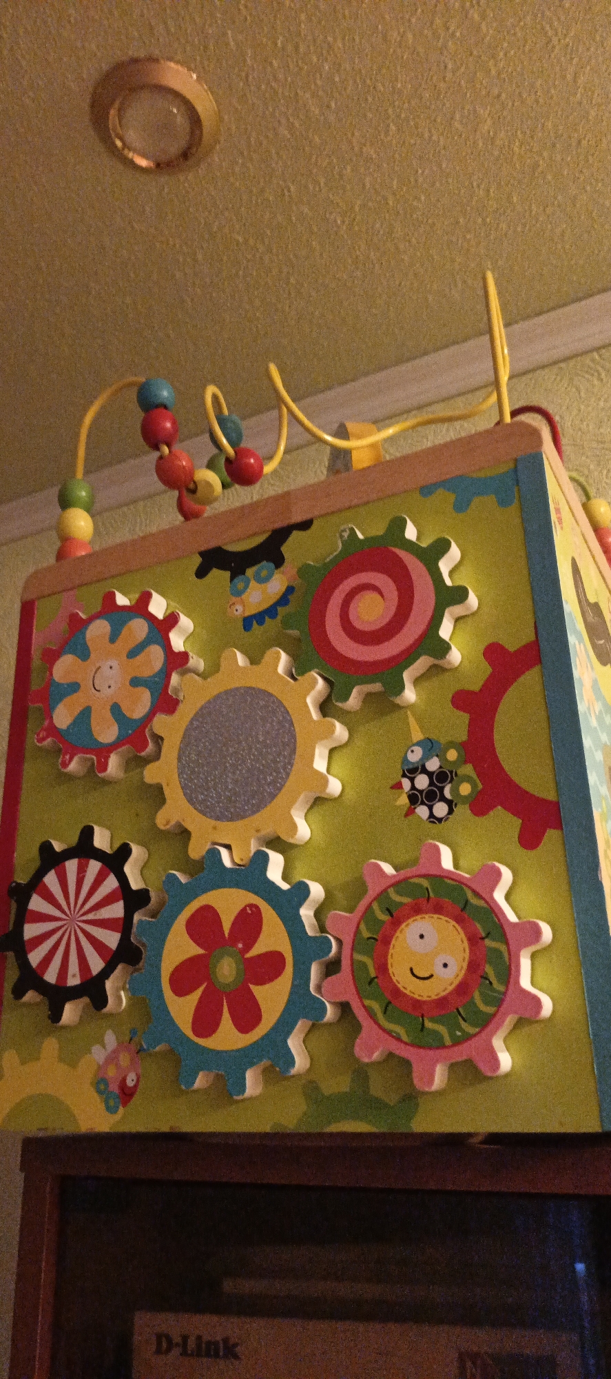 The image shows a colorful wooden activity cube for children, featuring various gears with different patterns and cartoon faces, as well as a bead maze on top. The cube is positioned on a dark wooden surface, possibly a shelf or table, which partially obscures a device labeled "D-Link" below. The background consists of a textured, light yellow ceiling with a circular light fixture, and a pale green, textured wall. The overall scene appears to be indoors, likely in a residential setting. Based on the provided information, this scene is located in Tikhvin, Russia.