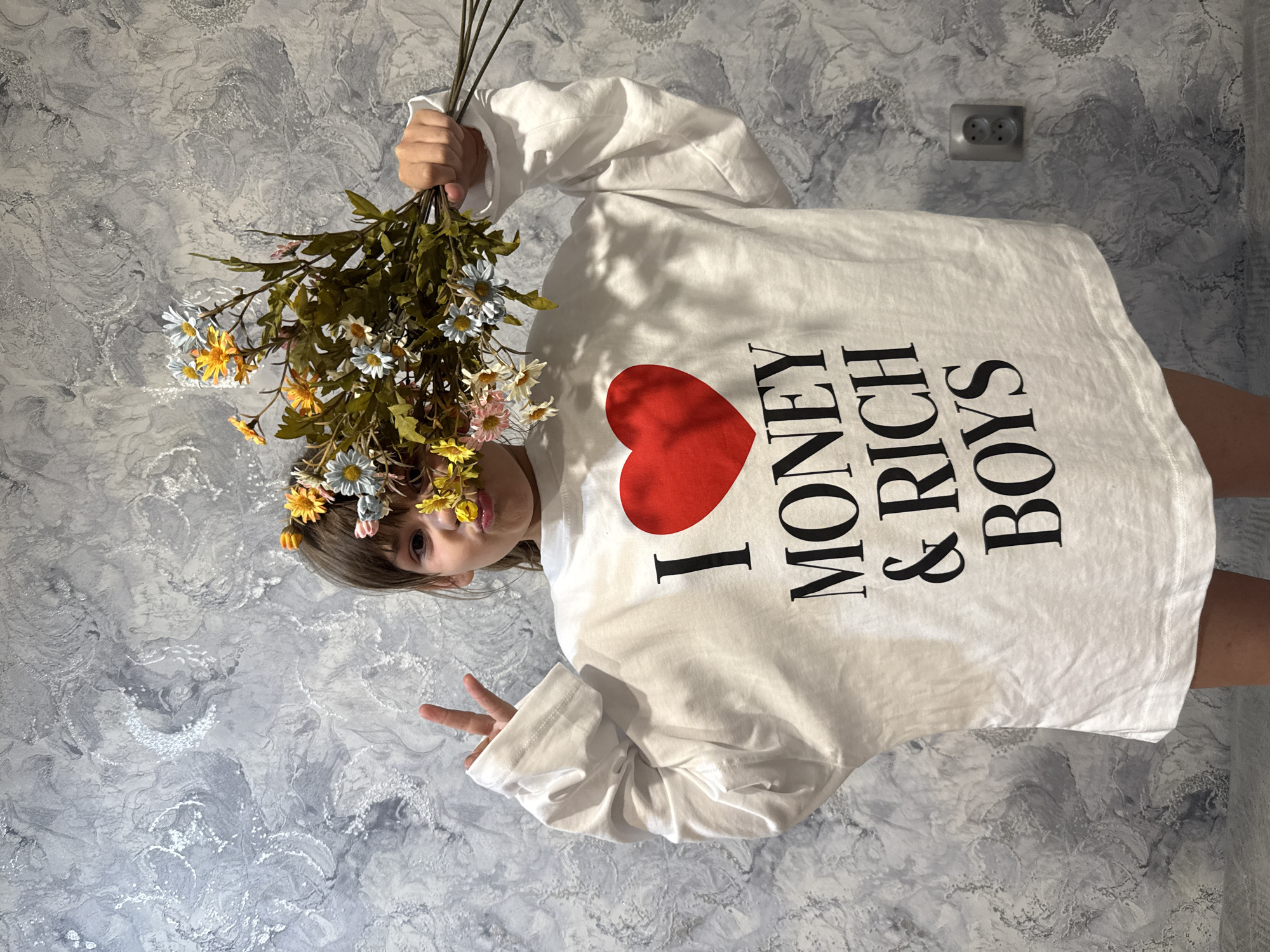 A young person with flowers in their hair is holding a bouquet of daisies. They are wearing a white long-sleeve shirt with "I (heart) MONEY & RICH BOYS" printed on it. The background is a marble-patterned wall with an electrical outlet. The image is rotated. Rusanovo, Russia is not ascertainable.