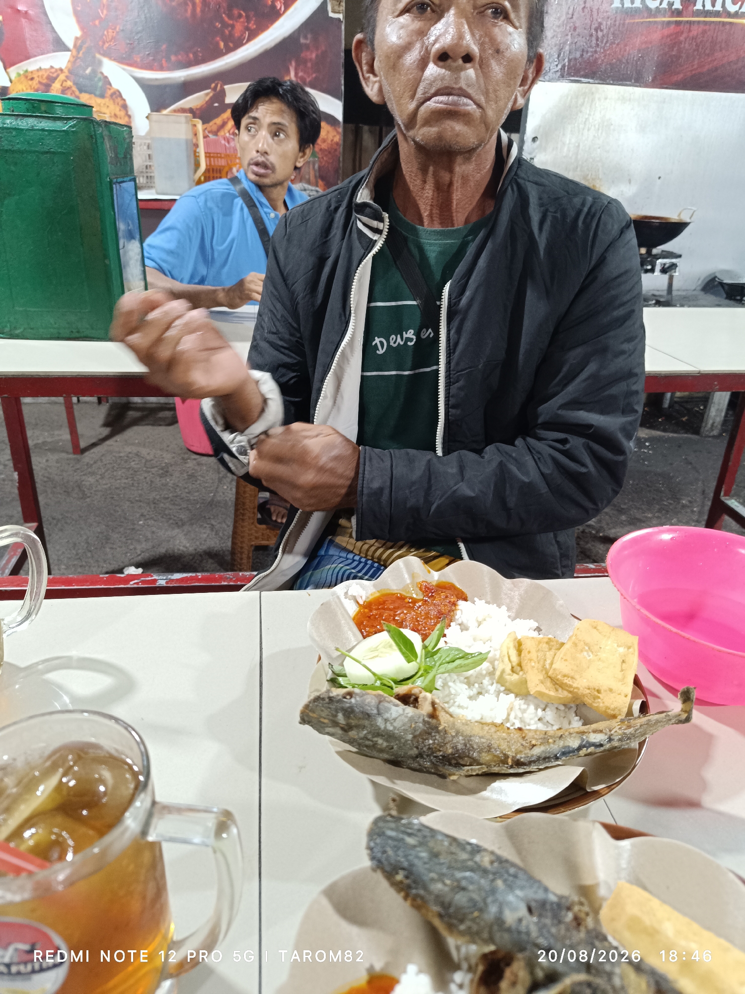 In Bringinbendo, Indonesia, a man in a black jacket is being filmed while eating a meal. The man in the foreground is looking forward, with his mouth slightly open, as if in conversation or about to speak. He has wrinkles on his neck and face, suggesting he is middle-aged or older. He wears a green t-shirt with white text that reads "Deus es". His hands are near his sleeves, and he appears to be adjusting his jacket.
To the left of the frame, another man in a blue polo shirt is visible, looking to his right. He has dark hair and a mustache. He is positioned behind a green container.
The meal consists of white rice, fried fish, fried tofu, cucumber slices, and a dollop of red sauce, served on brown paper. A glass of iced tea with a red straw is in the lower left corner. A pink bowl is situated on the right side of the table. The background shows a food stall with a large image of food on the wall and red tables and chairs. The timestamp "20/08/2026 18:46" is visible in the bottom right corner. The overall setting appears to be an outdoor eatery or food stall at night.