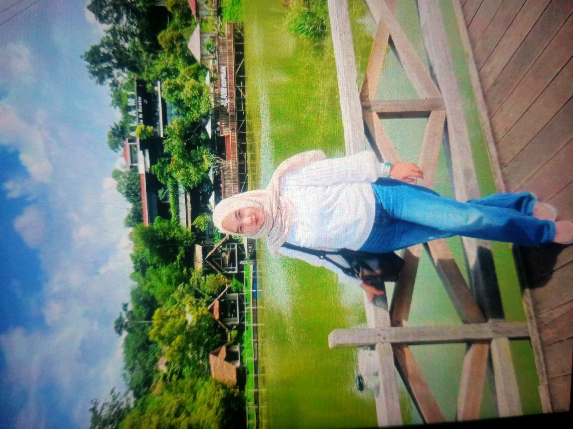 A young woman wearing a light pink hijab, a white long-sleeved shirt, and blue jeans stands on a wooden bridge. She is partially visible from the shoulders up, looking towards the camera with a slight smile. She has a black bag slung across her body. The bridge is made of weathered wooden planks and railings, with a green body of water visible below and behind her. In the background, there are lush green trees and some wooden structures, possibly huts or restaurants, on the edge of the water. The sky is blue with white clouds. The scene appears to be a scenic outdoor location. The image is rotated 90 degrees counter-clockwise. Based on the context provided, the location is Nusa Agung, Indonesia.