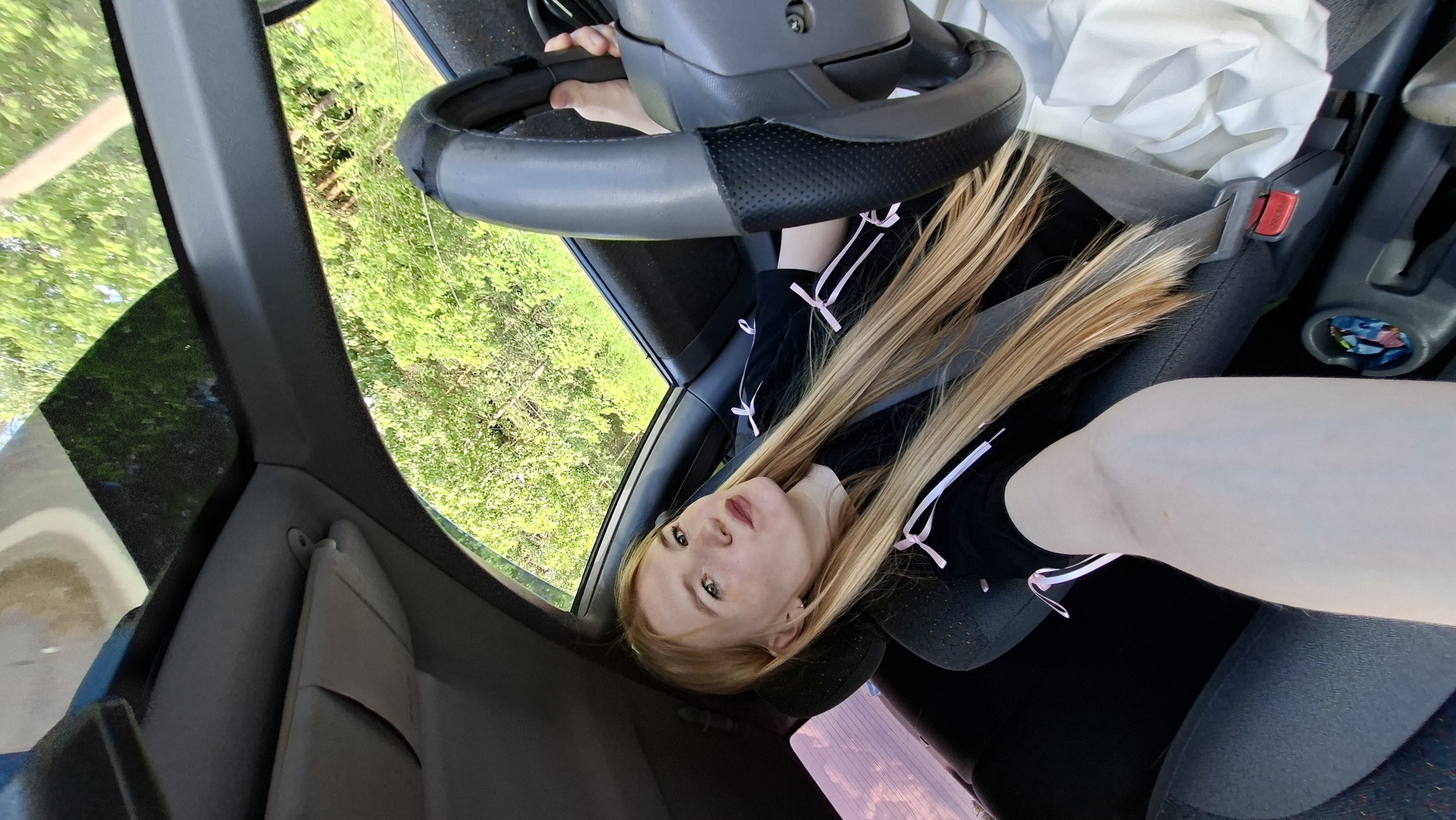 A young Caucasian woman with long blonde hair is in the driver's seat of a car, holding the steering wheel. She is wearing a black shirt with pink bows. The car's interior is visible, including a sunroof and a gray seatbelt. Outside the car, lush green trees are visible through the window. The image appears to be taken in a rural or suburban area, possibly near Chaikovsky, Russia, given the typical vegetation.
