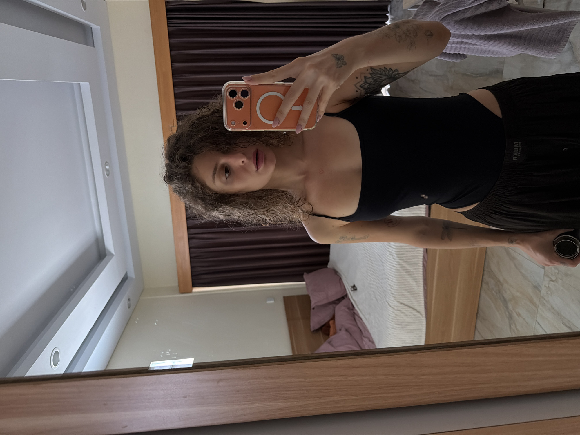 In a bedroom in North Kuta, Indonesia, a woman with curly brown hair is taking a mirror selfie. She is wearing a black tank top and dark brown pants. She holds an orange smartphone in her right hand, which is capturing the reflection. In the background, a bed with pink pillows is visible, along with dark brown curtains and a wooden wardrobe. The floor is tiled with a marble pattern.