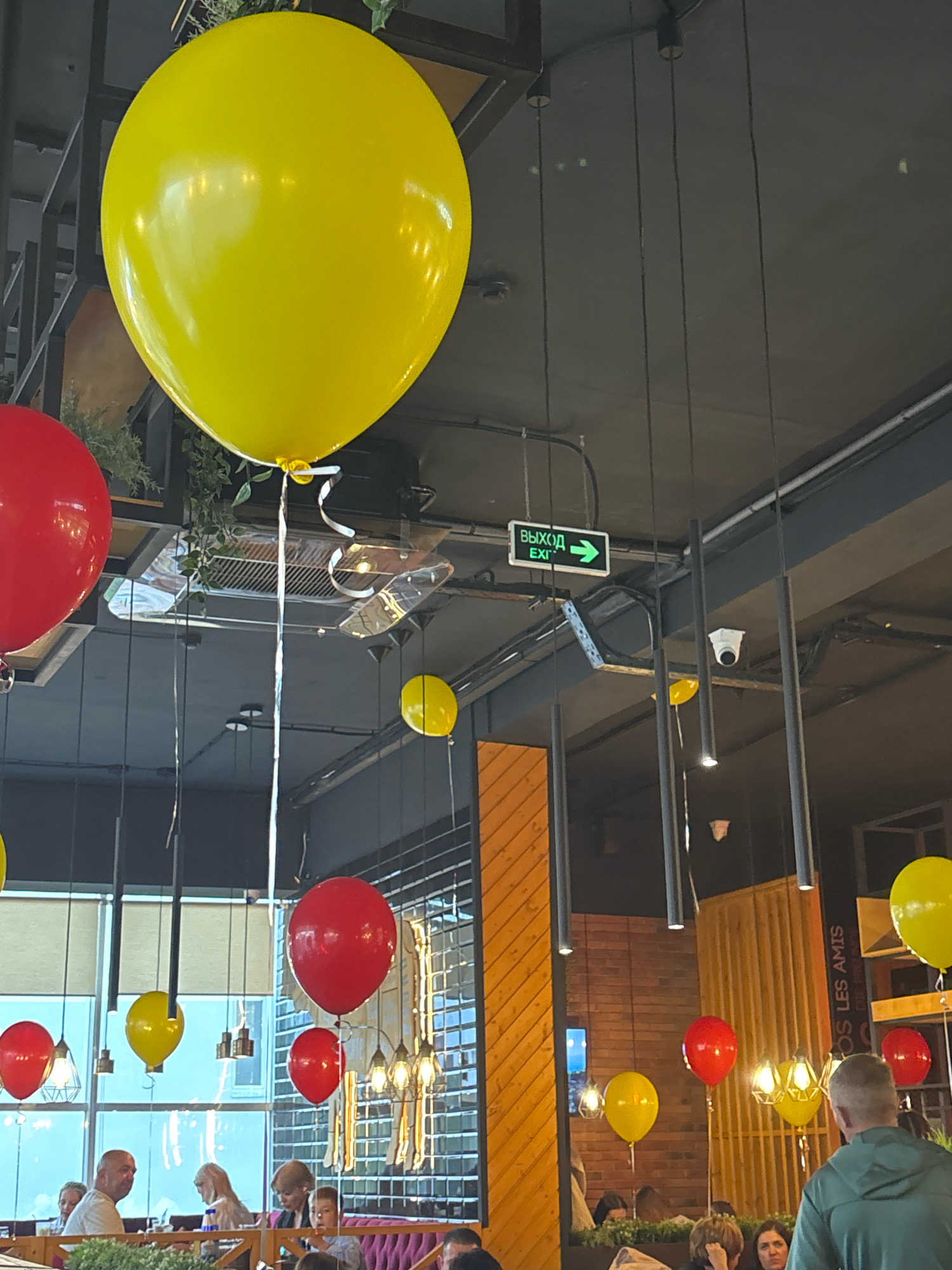 The image appears to be taken inside a brightly lit, modern establishment, possibly a restaurant or cafe, judging by the tables and seating arrangements visible in the lower portion of the frame. The ceiling features industrial-style exposed beams and ductwork, with several contemporary pendant lights and a security camera. A prominent green exit sign with Cyrillic text is affixed to the ceiling, indicating "ВЫХОД" (Exit) with a right-pointing arrow.
The environment is decorated with numerous colorful balloons, primarily in red and yellow, suspended from the ceiling by thin strings. Some of these balloons are quite large, while others are smaller. There are also large windows that provide a view of the outdoors, suggesting it is daytime.
Several people are visible in the foreground and mid-ground, seated at tables. They appear to be engaged in conversation and dining. Their clothing is casual.
The presence of the Cyrillic exit sign strongly suggests the location is within Russia. Without specific visual cues linking it to Blagoveshchensk, a definitive location within that city cannot be confirmed. However, the overall aesthetic is consistent with a modern urban commercial space in Russia.