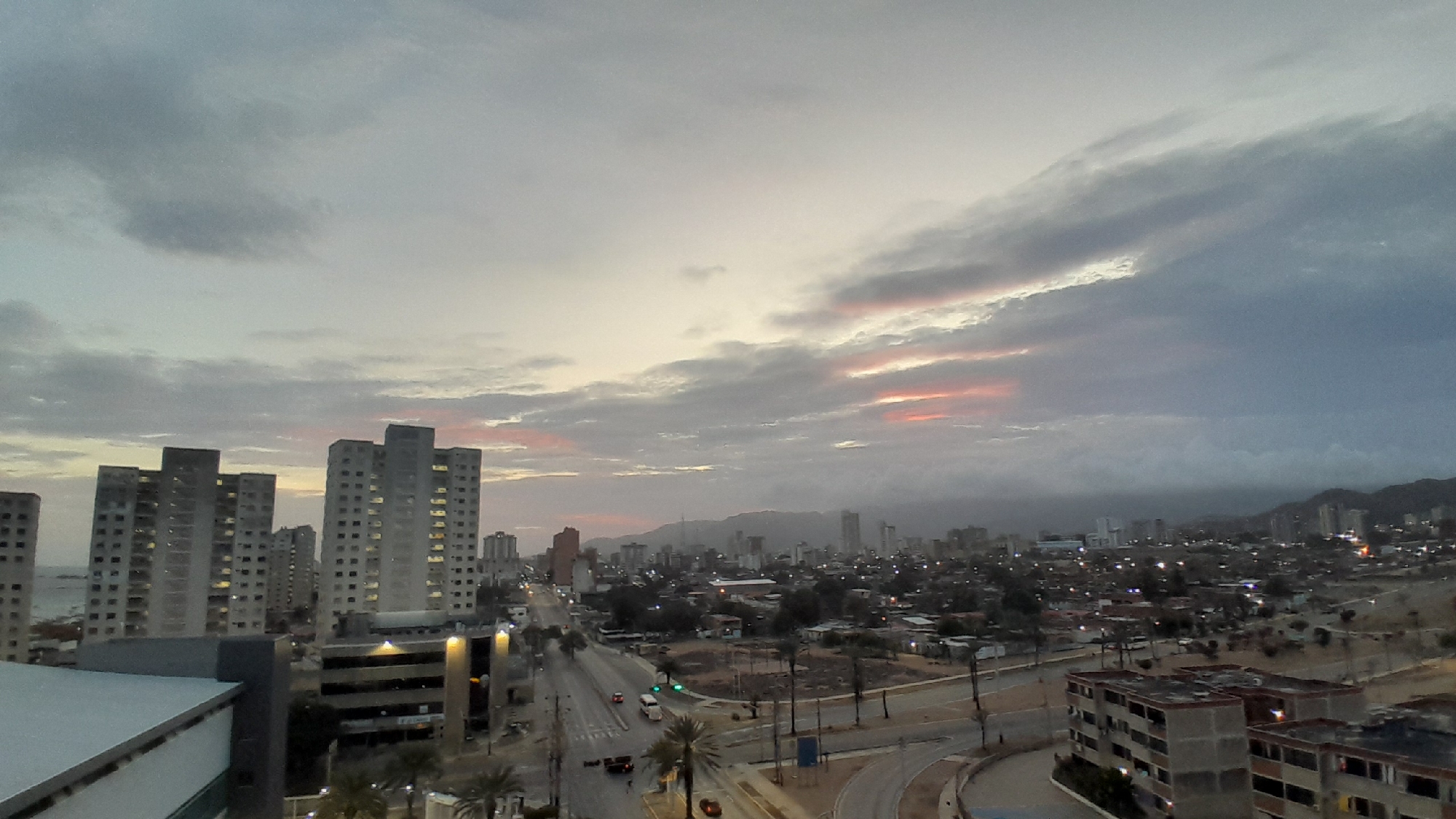 This is a wide-angle, eye-level outdoor photograph of a cityscape at dusk in Porlamar, Venezuela. The sky is filled with dramatic clouds that range from dark gray to lighter shades, with streaks of pink and orange indicating the setting sun. In the foreground on the left, a large, modern building with a flat roof and concrete facade is visible. To the right and slightly behind it are several tall, residential high-rise buildings with numerous windows, many of which are illuminated. A multi-level parking structure with visible lights is situated in front of these buildings.
Further into the city, a wide, multi-lane road runs through the center of the frame, with several vehicles, including a white bus and a red car, visible on it. Streetlights are beginning to illuminate the road and surrounding areas. Palm trees are scattered throughout the scene, particularly along the roadside and near the residential buildings. In the mid-ground, a dense urban landscape with a mix of smaller buildings and more high-rises extends towards the background. The far background is dominated by a hazy mountain range that is partially obscured by the clouds. The overall atmosphere is one of a bustling city transitioning into night, with a dramatic sky providing a natural spectacle.