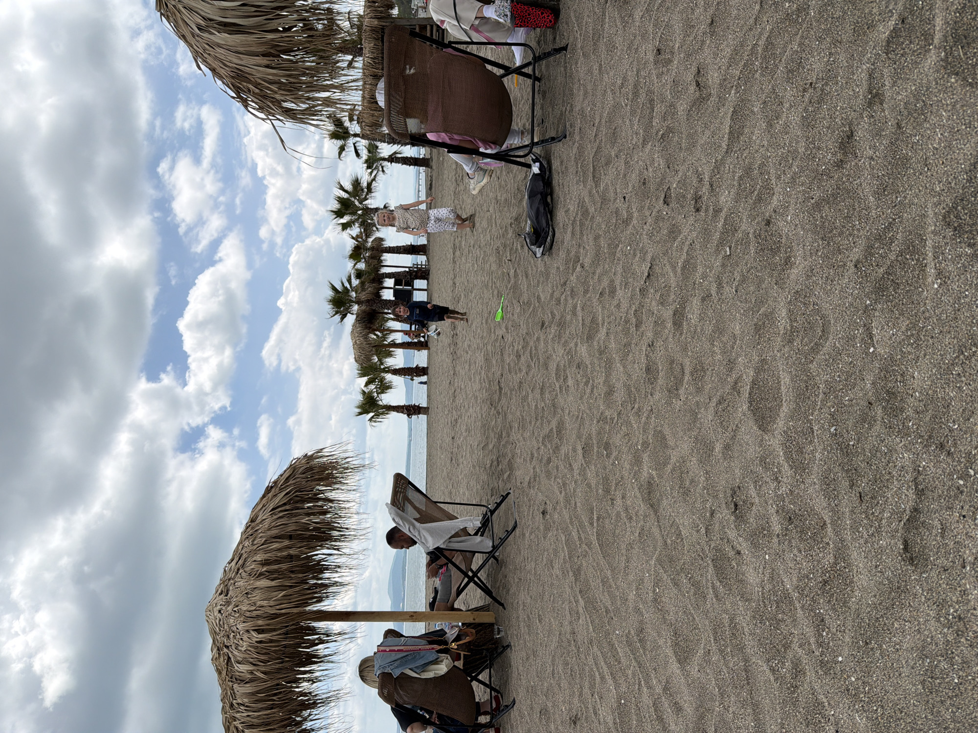 The image shows a beach scene with dark sand, palm trees, and people relaxing. The location is Unknown, Unknown.
People are seated in beach chairs under straw umbrellas. Children are present, standing and looking towards the camera. A black bag is on the sand near a chair. The sky is cloudy, with patches of blue.