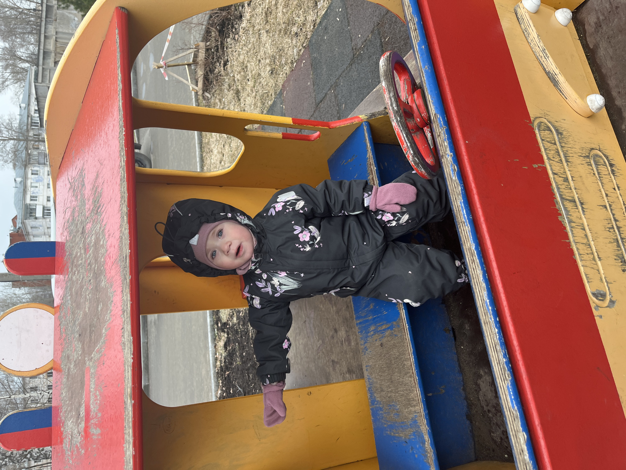 A young child in a black snowsuit with pink gloves and a matching hat is situated inside a colorful playground structure, likely in Chaikovsky, Russia. The structure features red, yellow, and blue paint, with a red steering wheel visible near the child. The playground is outdoors, with patches of dirt and grass suggesting a late winter or early spring setting. The child is looking up and seems to be in good spirits. The background contains buildings, possibly residential.