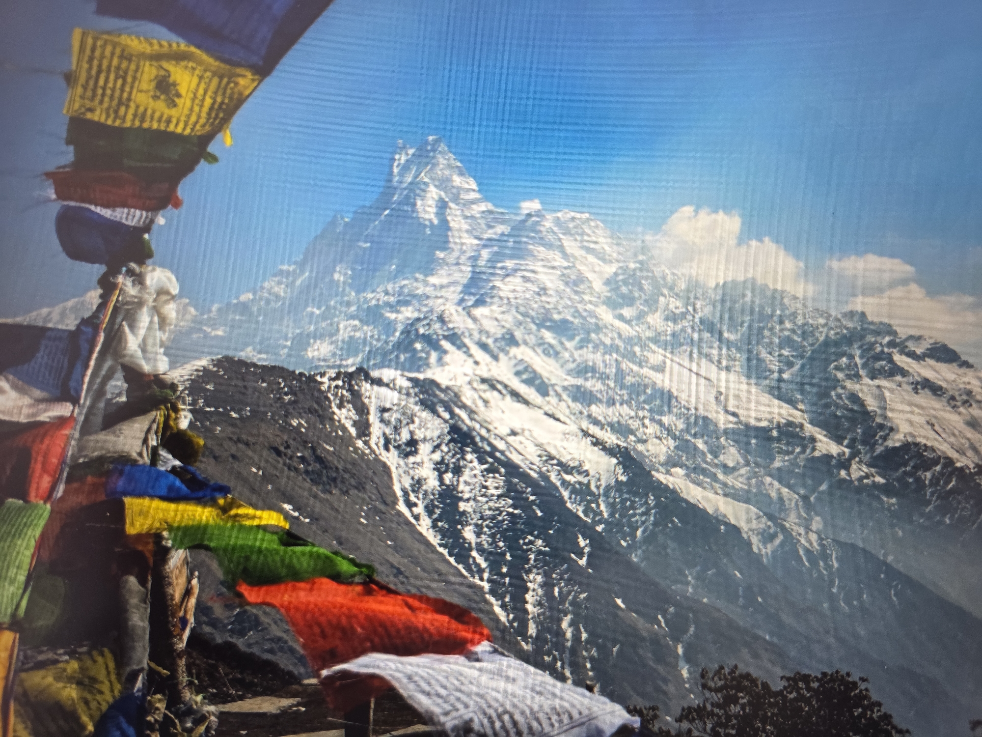This media file depicts a majestic mountain landscape under a bright, clear blue sky, with some scattered clouds. In the foreground, colorful prayer flags are strung up, fluttering in the wind. The flags are predominantly blue, yellow, green, red, and white, some appearing to have symbolic imagery or text printed on them, though not clearly legible. These flags are attached to wooden poles and are a prominent visual element on the left side of the image.
Behind the flags, the terrain slopes upwards, revealing a rugged, rocky mountainside with patches of snow. The dominant feature of the scene is a towering, snow-capped mountain peak that rises dramatically in the distance. The mountain appears sharp and imposing, with deep shadows and stark highlights accentuating its contours. The overall impression is one of natural grandeur and a serene, high-altitude environment. The sunlight suggests it is daytime, likely mid-morning or early afternoon, given the clear sky and the brightness of the scene. The presence of prayer flags suggests a spiritual or cultural context, often found in mountainous regions in South Asia. However, the provided location context of Atyrau, Kazakhstan, is inconsistent with the typical setting for such prayer flags and mountainous scenery. Therefore, while the image visually represents a mountainous scene with prayer flags, the specified location context cannot be directly inferred from the visual elements alone.