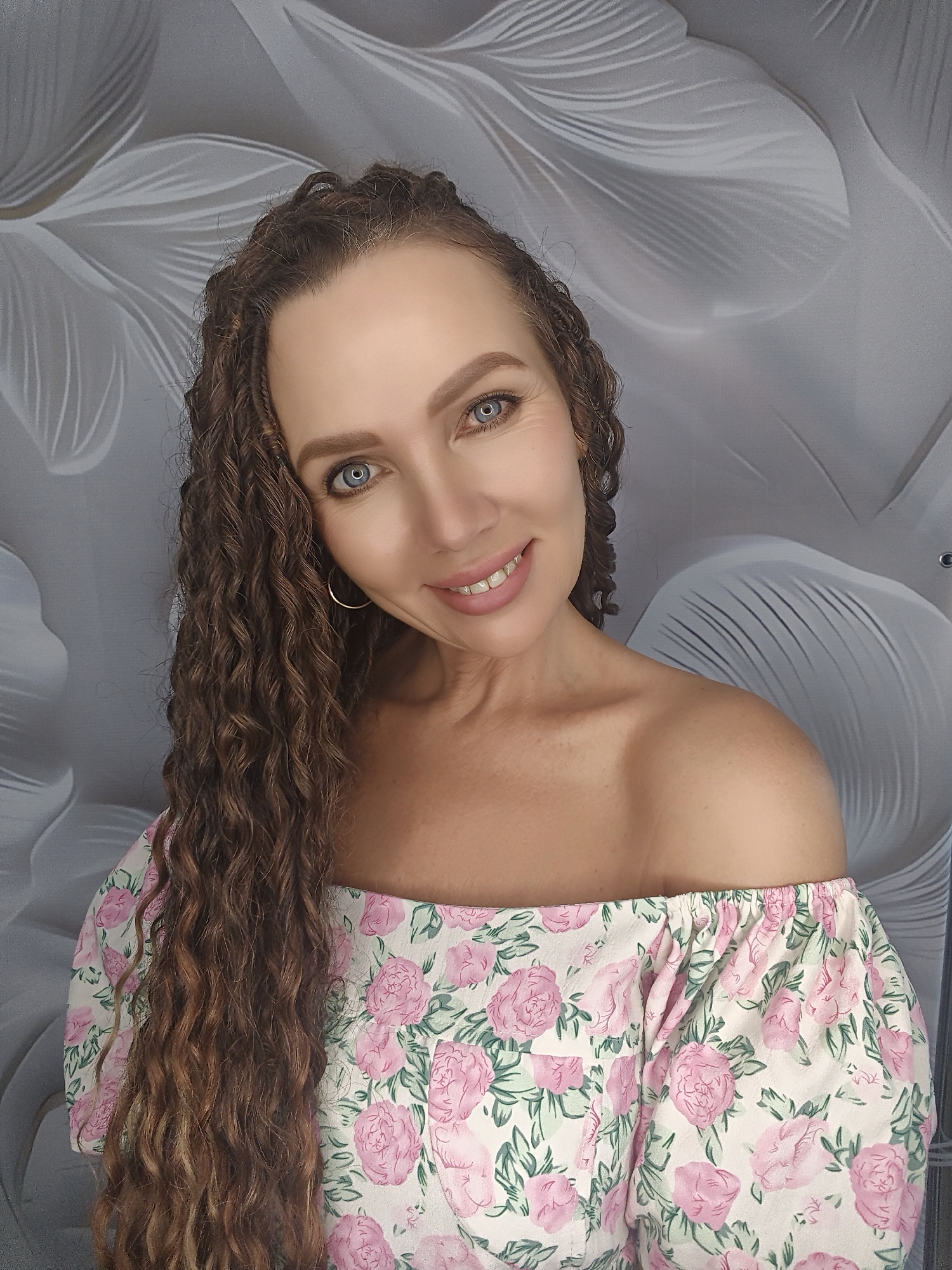 A woman with long, curly brown hair and striking blue eyes smiles at the camera. She is wearing an off-the-shoulder top with a floral pattern of pink roses and green leaves against a white background. Her hair cascades down over her left shoulder, and a delicate hoop earring is visible in her left ear. The background is a grey wall with a subtle, abstract floral design. The lighting appears to be soft and even, suggesting an indoor setting, possibly a studio or a well-lit room. Her expression is warm and inviting, conveying happiness and confidence. There is no visible text in the image.