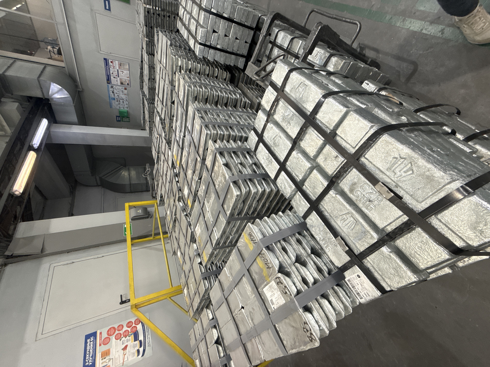 The image depicts stacks of strapped, rectangular aluminum ingots in an industrial setting. The stacks are arranged in multiple rows, and some are placed on metal carts or supports. The background reveals a portion of an industrial building, including ductwork, lighting fixtures, and informational posters on the walls. One person wearing athletic shoes is partially visible in the top right. The location is presumed to be in Aramil, Russia.