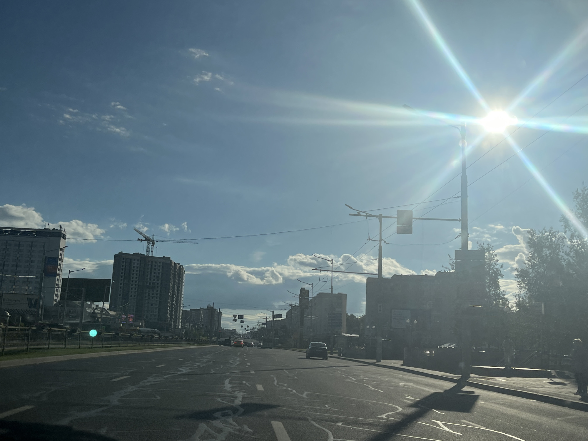 The image depicts a view of a city street in Minsk, Belarus, on a bright, sunny day. The sun is high in the sky and flares brightly, creating a lens flare effect across the image. The sky is blue with scattered white clouds.
The street is wide and appears to be a multi-lane road with lane markings visible. There are several vehicles on the road, including cars and what appears to be a bus in the distance. Traffic lights and streetlights are present along the roadside.
Buildings of varying heights line the street. Some appear to be residential or office buildings, while others are under construction, indicated by the presence of a crane in the background. Trees and foliage are also visible along the sidewalks and in some areas between buildings.
The overall scene suggests a typical urban environment, possibly in a busy part of the city. The weather appears pleasant, characteristic of a sunny day. The perspective is from within a vehicle, suggesting the image was taken while driving.