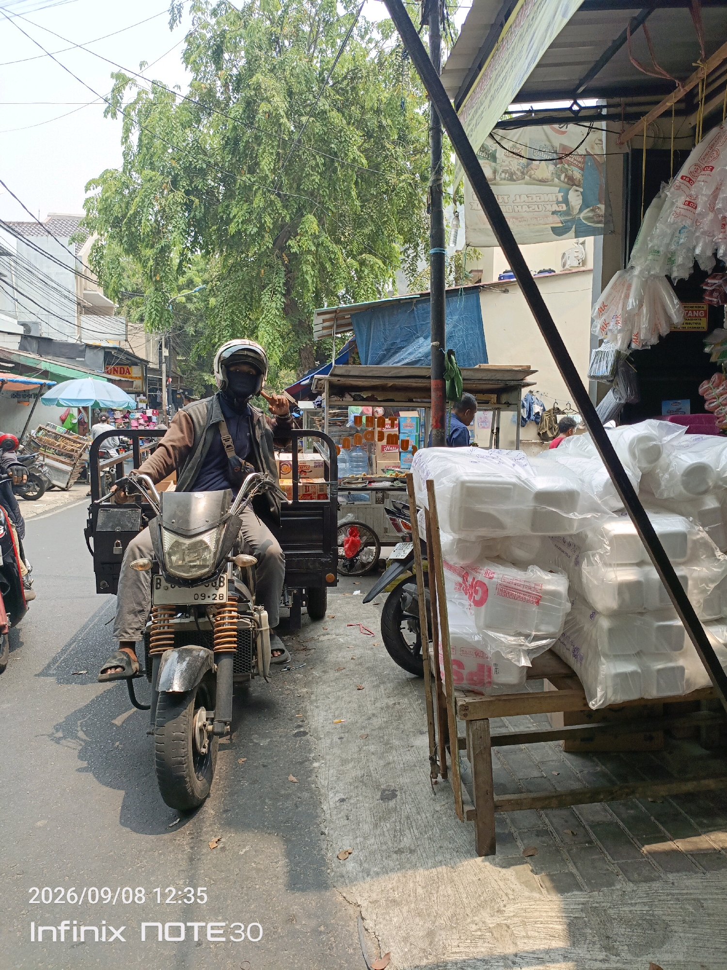 A man wearing a helmet and a face mask is riding a three-wheeled motorcycle down a street in Central Jakarta, Indonesia. The motorcycle has a cargo area behind the rider, filled with white styrofoam containers. The man is wearing a brown jacket, a blue shirt, and gray pants. He is holding onto the handlebars and has his left index finger raised, possibly signaling. The street is paved with asphalt and lined with buildings and trees. To the right of the motorcycle, there is a stall selling packaged goods and more styrofoam containers. The sky is bright and clear. The timestamp on the image indicates it was taken on September 8, 2026, at 12:35 PM.