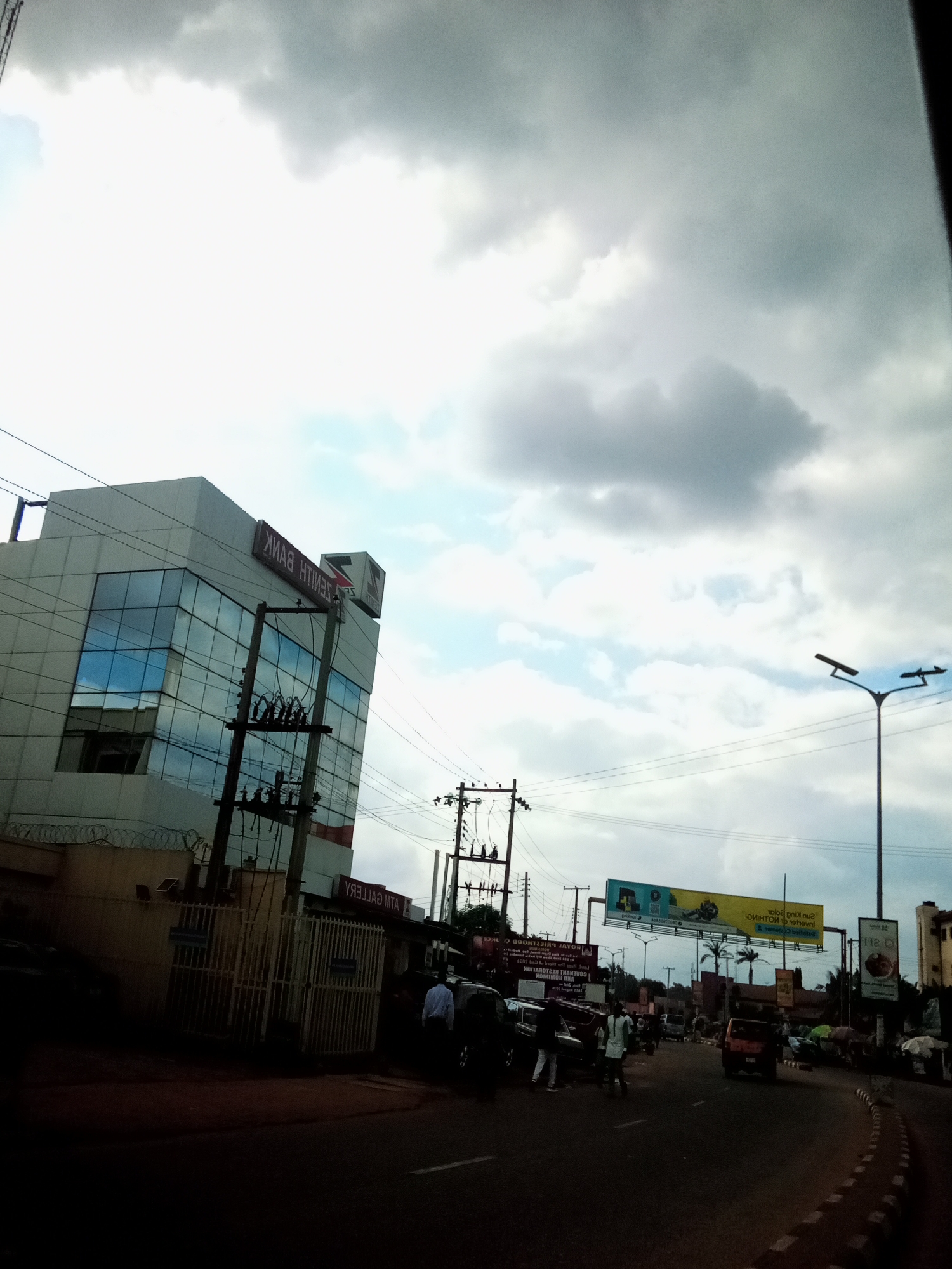 The image shows a street scene in Nibo, Nigeria. A modern, multi-story building with reflective glass windows is prominently featured on the left side of the frame. The building has "ZENITH BANK" signage visible on its facade. Electrical poles and overhead wires are present, some appearing to support streetlights. The street itself is paved and appears to be a busy thoroughfare with several vehicles, including cars and possibly a bus, traveling or parked along the roadside. Pedestrians can be seen on the sidewalk and near the vehicles. A large billboard advertising a solar product is suspended over the street in the middle ground. The sky is overcast with a mix of light and dark clouds, suggesting a cloudy or potentially rainy day. The overall environment depicts an urban setting with commercial activity.
