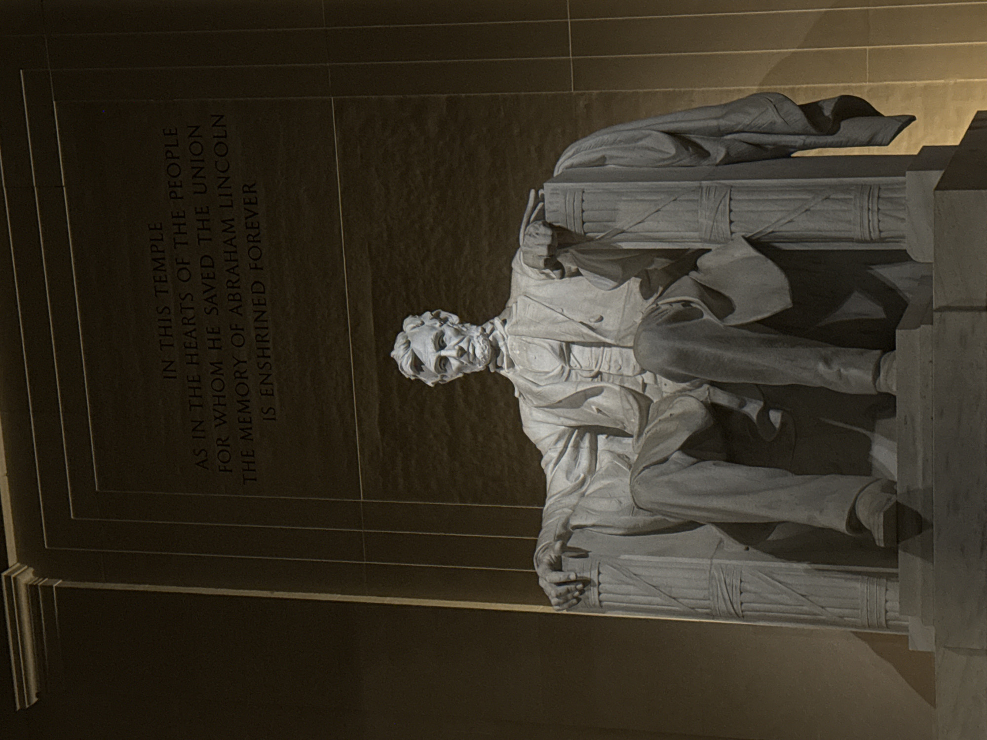 The image depicts the statue of Abraham Lincoln within the Lincoln Memorial, located in Washington, D.C., United States of America. The statue, carved in white marble, shows Lincoln seated, with a draped cloth behind him. Behind the statue is a wall inscribed with text about Lincoln's memory being enshrined forever. The lighting suggests an interior setting, possibly at night.