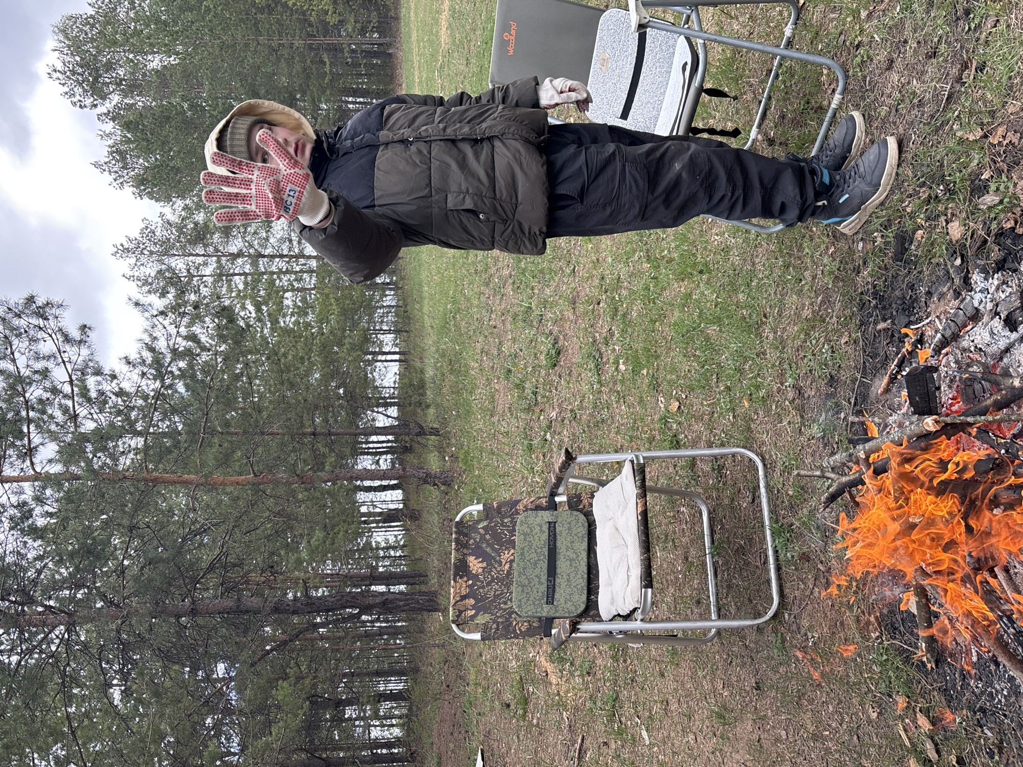 The image depicts an individual standing next to a campfire in an outdoor environment that appears to be Vekoshinka, Russia. The individual is wearing a hat, dark puffy jacket, dark pants and work gloves. They are holding up their gloved hand, palm facing forward, toward the camera. Behind the individual, there are pine trees and a grassy field. Two folding chairs, one with a camouflage pattern, are visible near the campfire and individual. The campfire has active flames with burning wood. The background shows more trees under a cloudy sky.