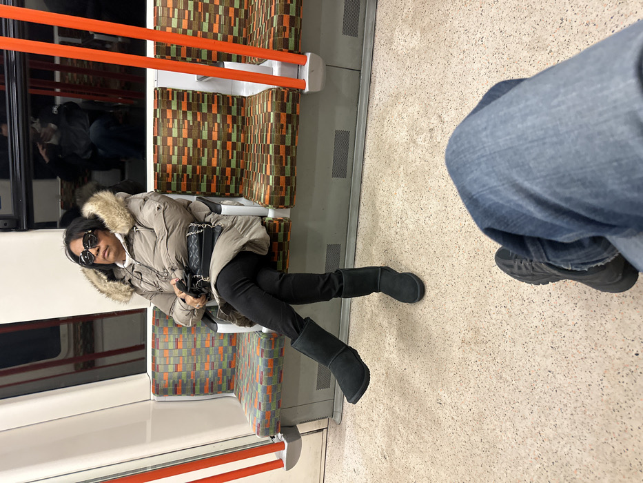 The image displays the interior of a London Underground train carriage. An adult female, seated on a patterned fabric bench seat, occupies the left side of the frame. She wears large, dark sunglasses, a light brown or beige fur-hooded puffer coat over a lighter garment, black trousers, and black mid-calf boots. Her legs are crossed at the ankles, with her left foot elevated. She holds a black quilted shoulder bag with a silver chain strap in her hands. Her posture is upright, and her expression is neutral. The seats feature a distinctive geometric moquette pattern of orange, green, brown, and black squares. Vertical orange handrails are visible on the right side of the carriage. The floor is light grey and speckled. A reflection of an individual is visible in the darkened window panel above the seats on the right. In the immediate foreground, the knees and lower legs of a second individual, wearing blue denim jeans and dark athletic shoes, are visible, indicating the perspective from which the photograph was taken.