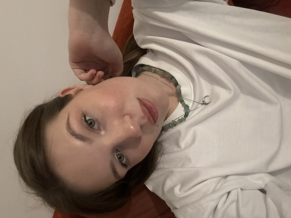 A close-up indoor portrait captures a young adult female with light brown hair and light-colored eyes, her head tilted slightly while looking directly at the camera. She is wearing a plain white long-sleeved crew-neck top. Around her neck are two necklaces: one composed of irregular green beads, and a second, finer silver-colored chain with a crescent moon pendant. Her right hand is positioned near her right ear. The background consists of a light-colored wall or surface behind her head, and a textured reddish-orange surface (possibly upholstery or a cushion) to her left and behind her right arm. The subject appears to be reclining. The location is Vozdovac Urban Municipality, Serbia.